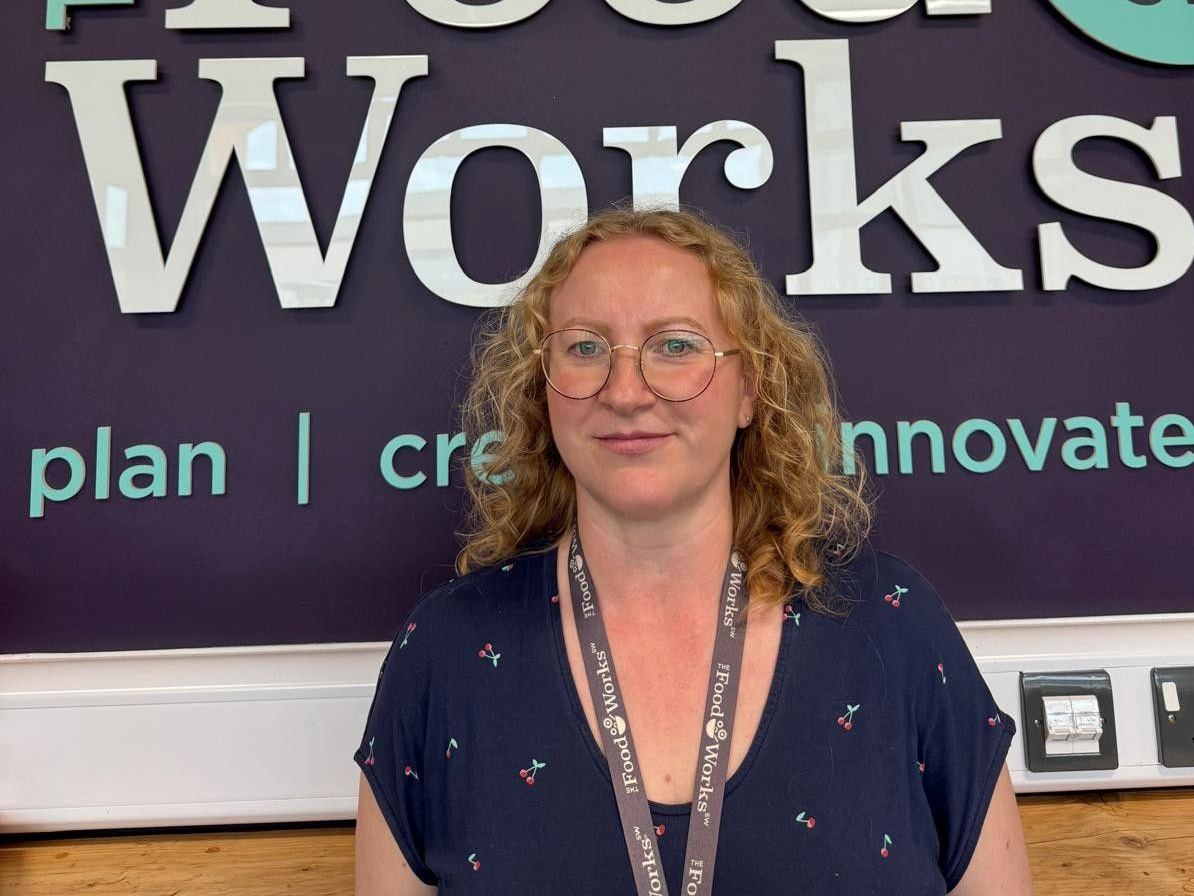 Woman with blonde curly hair and glasses standing in front of a sign that says 'Food Works'.