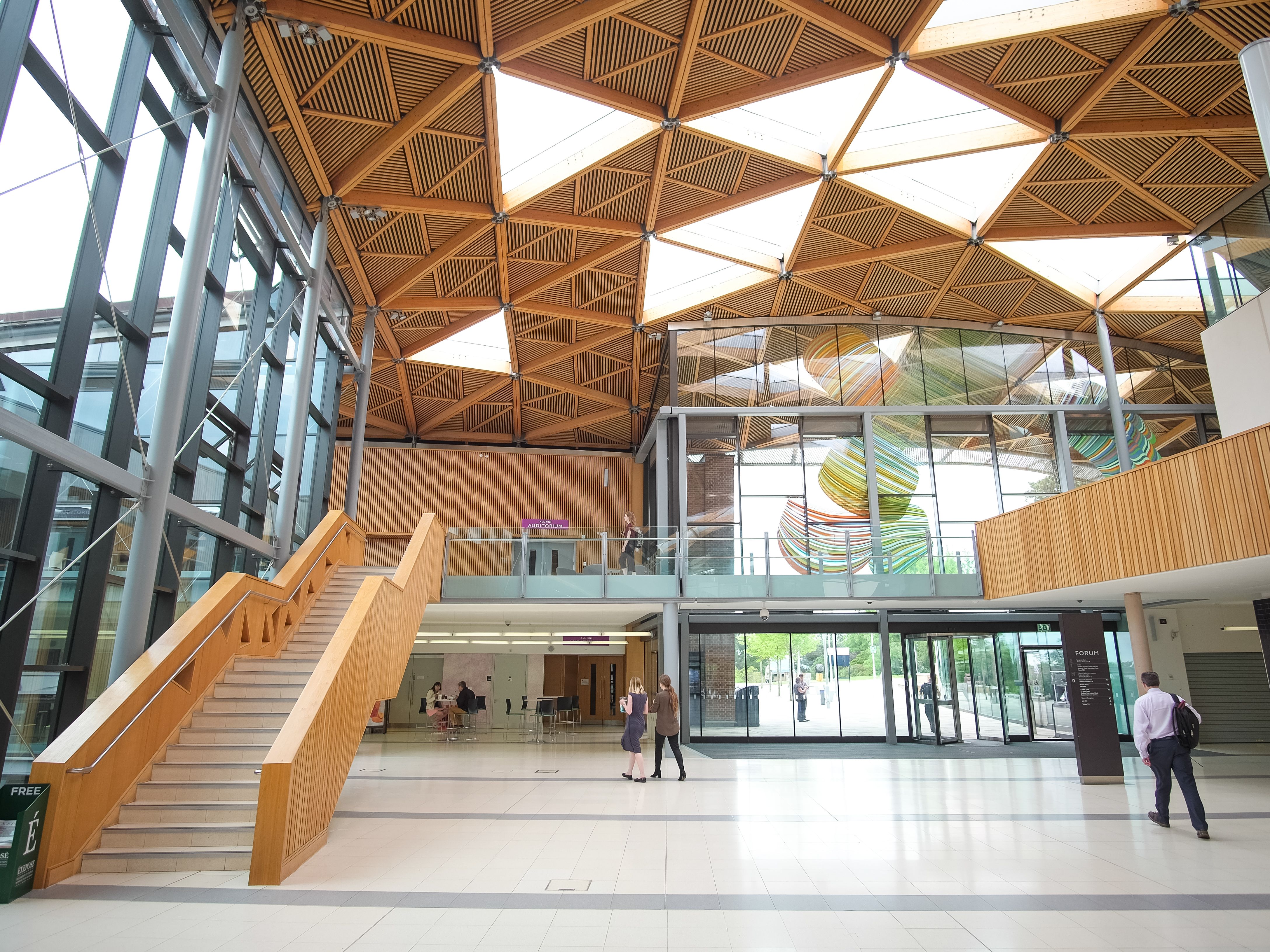 Modern indoor atrium with wooden geometric ceiling, glass walls, staircase, and people walking.