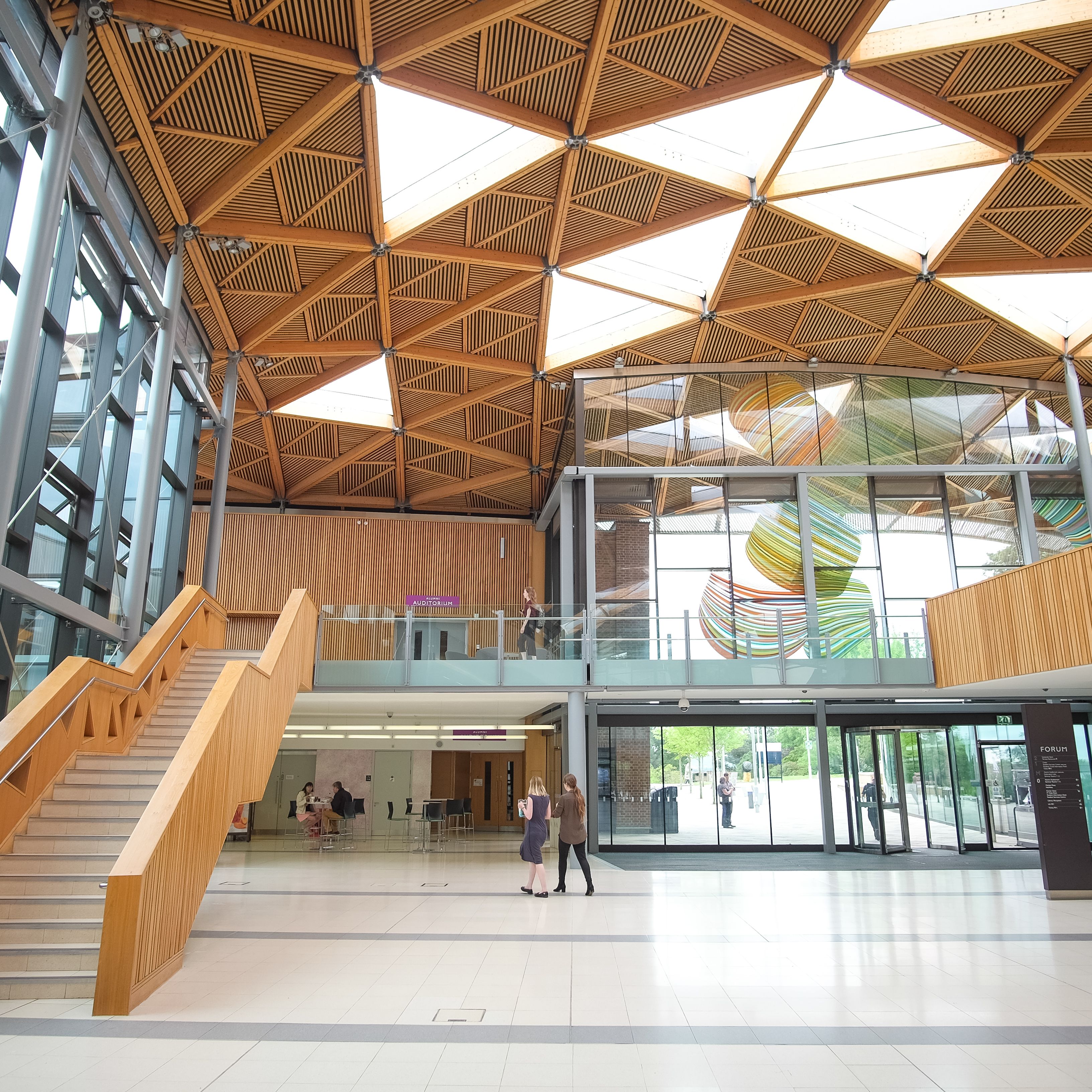 Modern indoor atrium with wooden geometric ceiling, glass walls, staircase, and people walking.