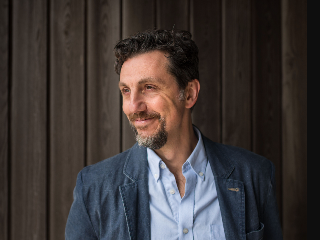Man with curly hair and goatee in a blue blazer and shirt, standing in front of a wooden wall.