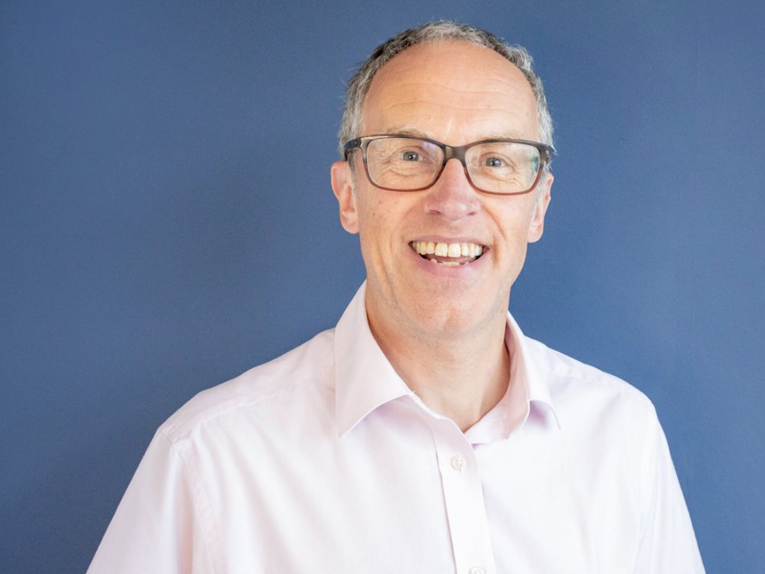 Smiling man wearing glasses and a light pink shirt standing in front of a blue background
