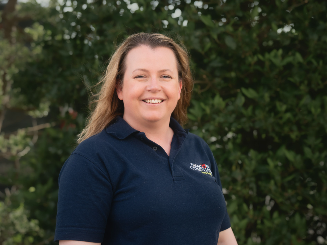 Smiling woman with long hair wearing a navy blue polo shirt standing outdoors in front of green foliage.