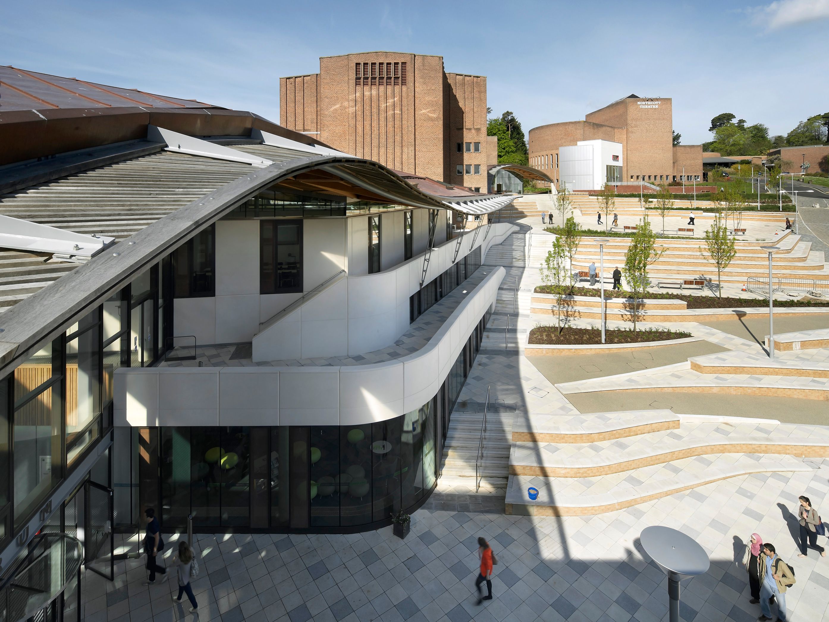 Modern university campus with distinctive brick and white buildings, open plaza, and landscaped terraces under a blue sky.