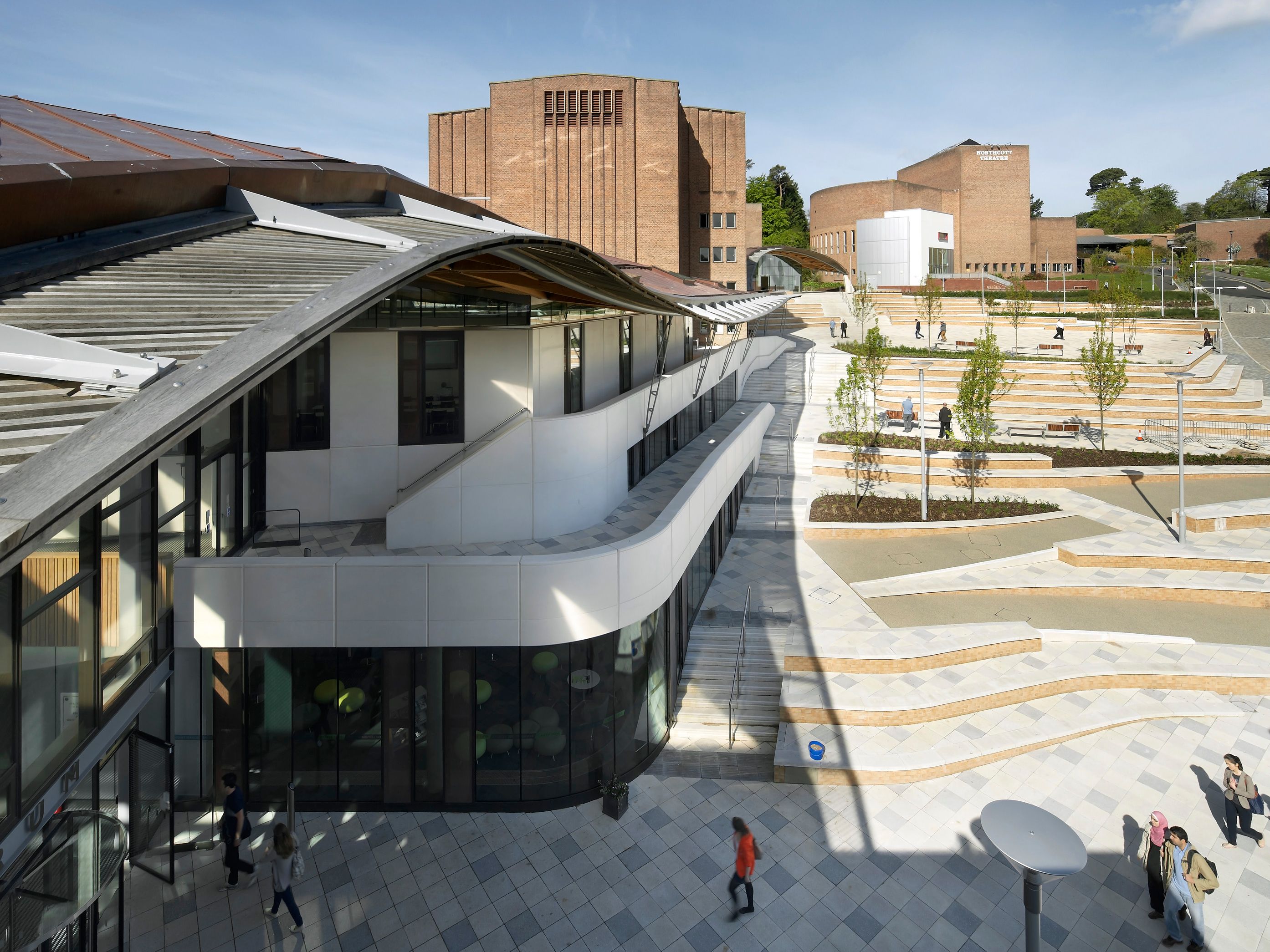 Modern university campus with distinctive brick and white buildings, open plaza, and landscaped terraces under a blue sky.