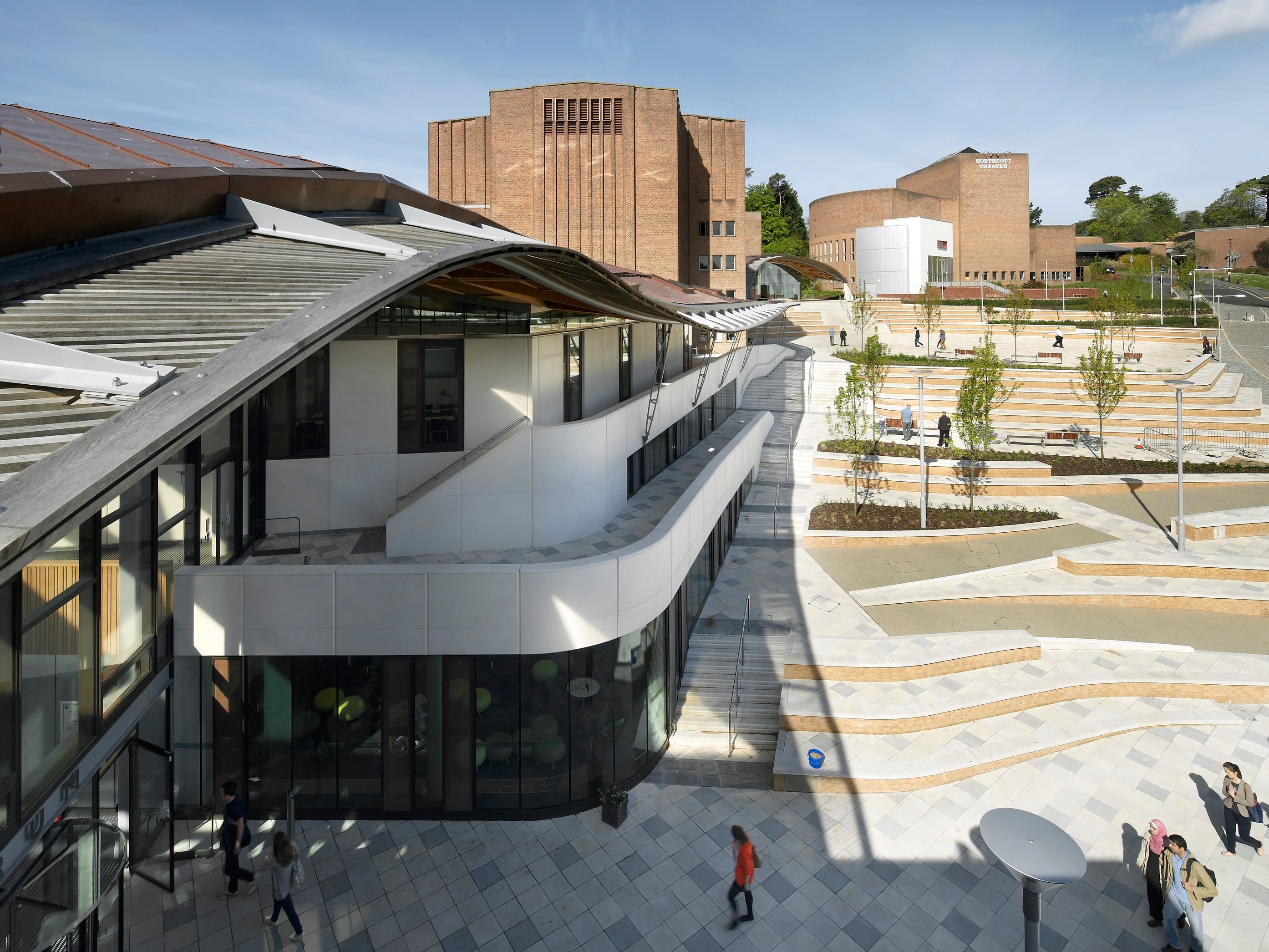 Modern university campus with distinctive brick and white buildings, open plaza, and landscaped terraces under a blue sky.
