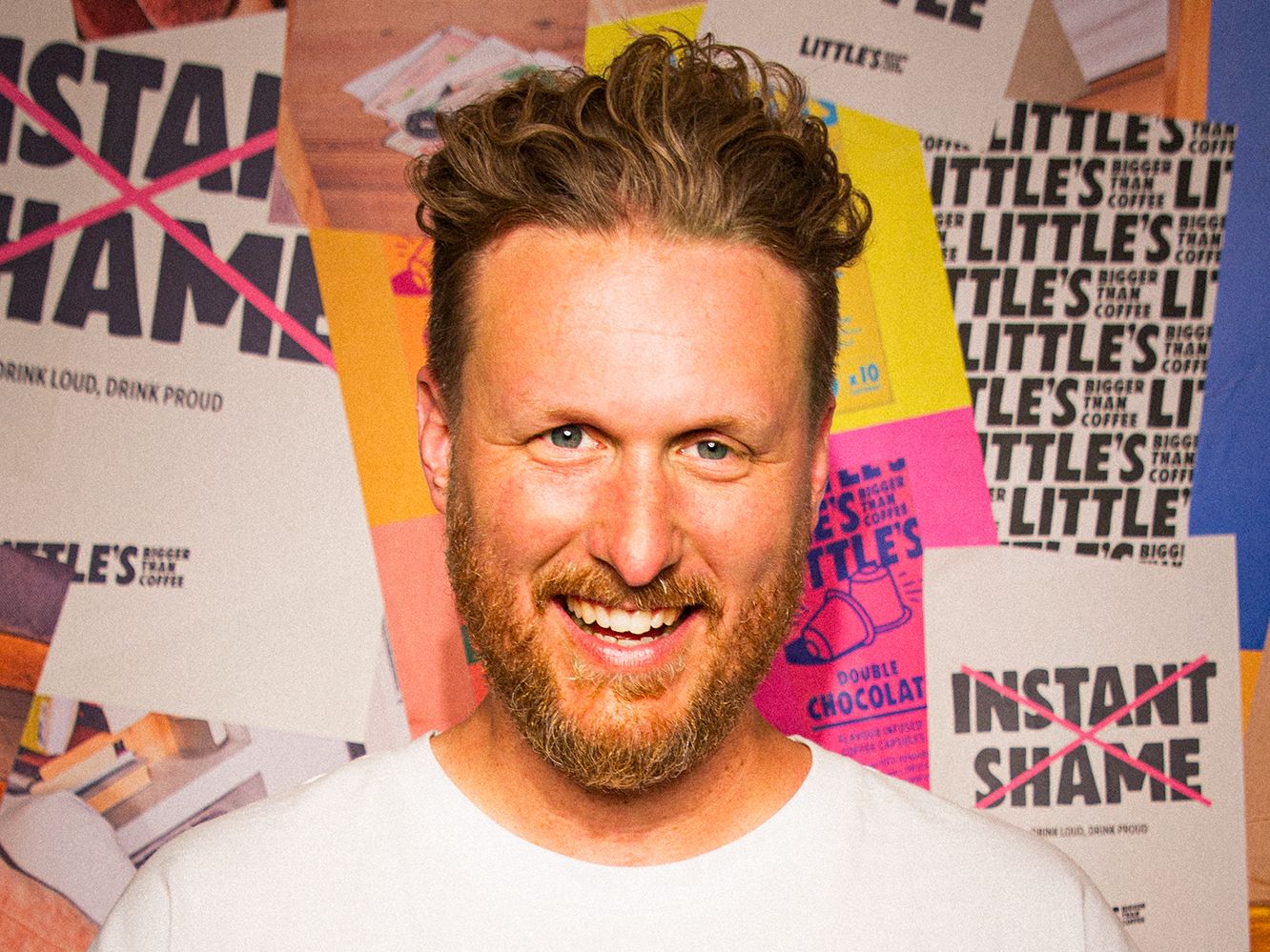 Smiling man with brown hair and beard wearing a white t-shirt standing in front of a colorful poster-covered wall.