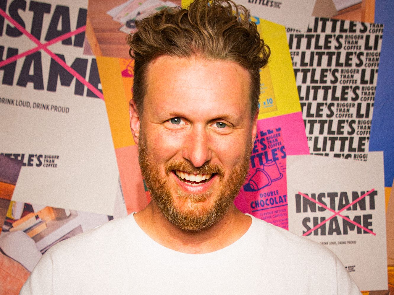 Smiling man with brown hair and beard wearing a white t-shirt standing in front of a colorful poster-covered wall.