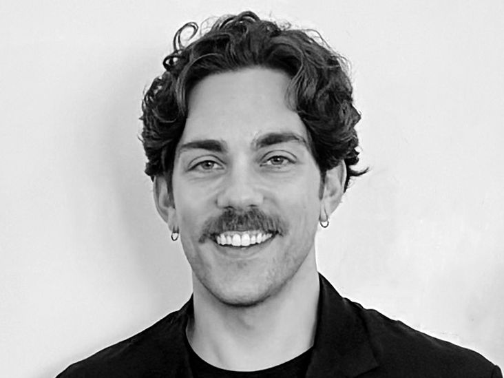 Smiling man with curly hair, mustache, and earrings in a black jacket