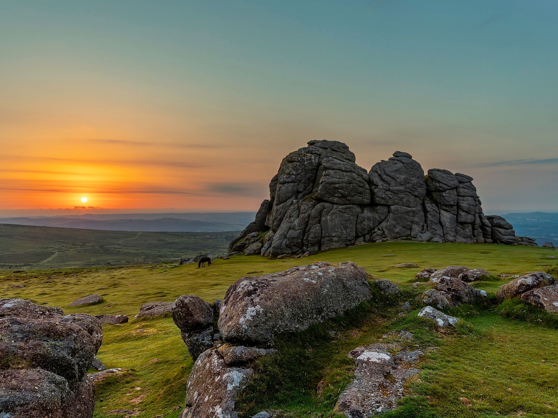 A serene sunset scene over Haytor with large stone formations and green grass. A lone horse grazes near the rocks as the sky transitions from golden hues near the horizon to a soft blue above