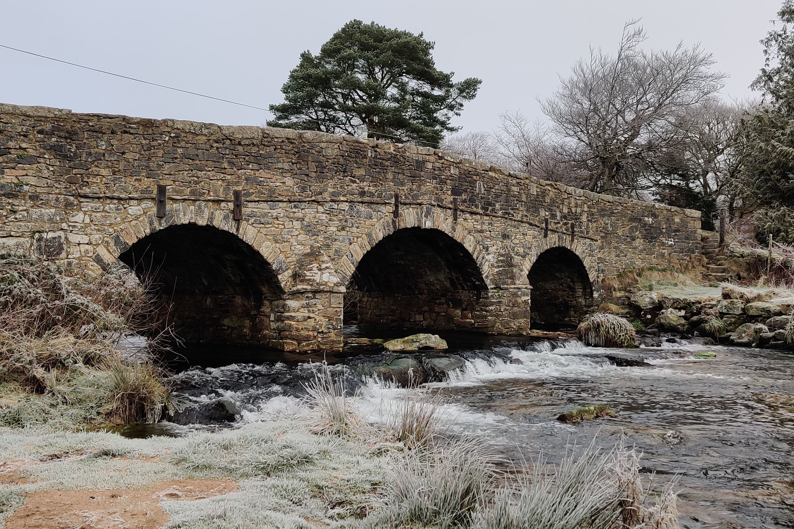 A rustic stone bridge with three arches spans over a flowing stream in a frosty, winter landscape. Bare trees and light frost on the grass add a tranquil, chilly atmosphere to the scene.