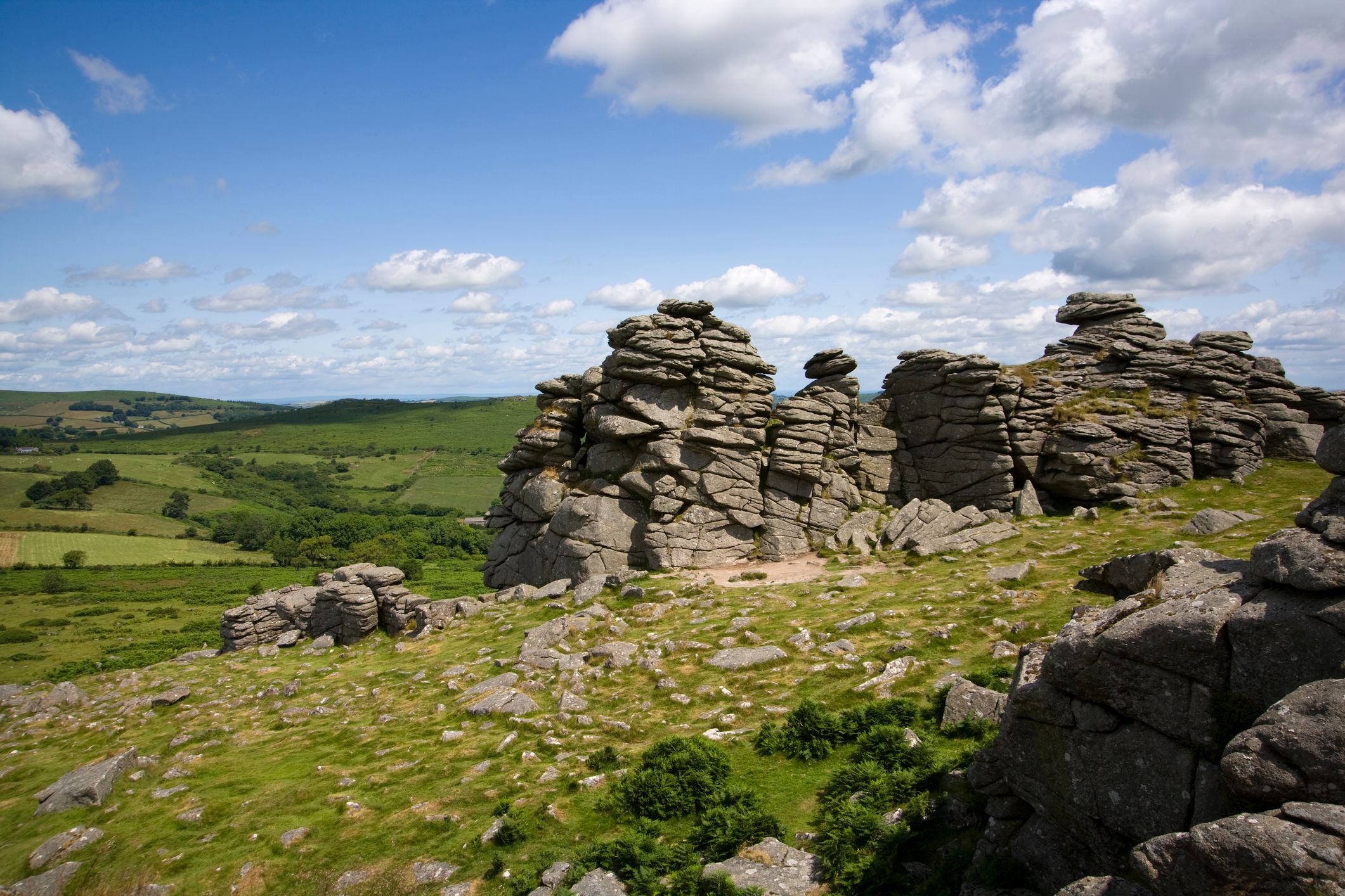 Houndtor sits atop a grassy hill under a bright blue sky dotted with fluffy white clouds. Rolling green fields and distant hills stretch out in the background.