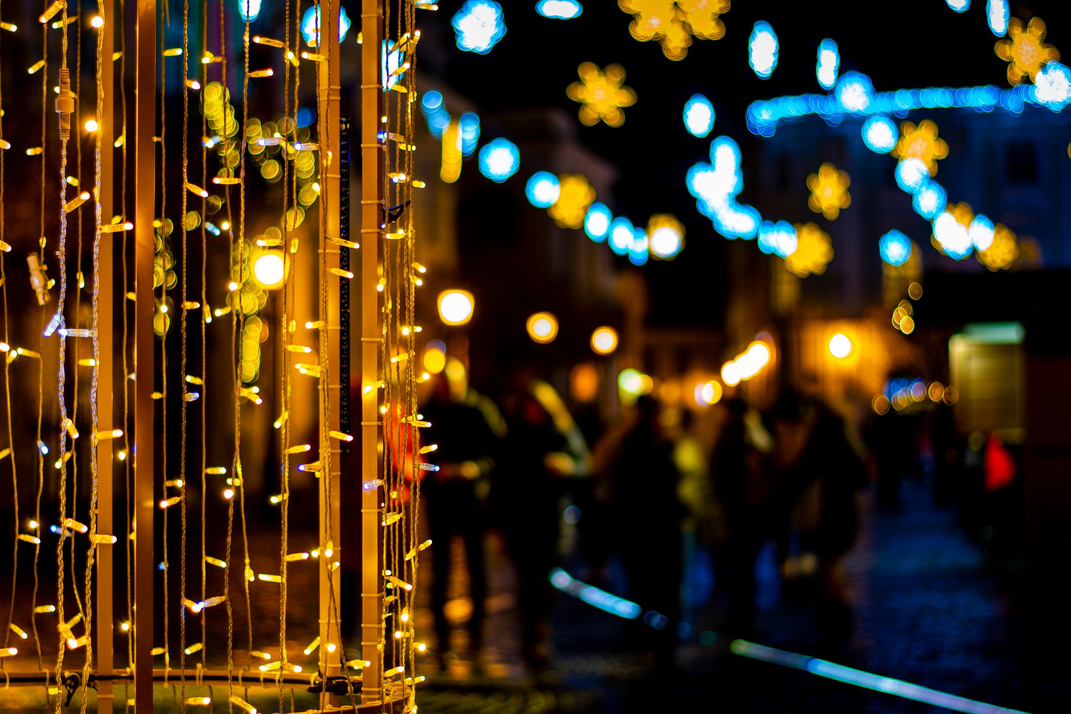 Twinkling string lights and glowing snowflake decorations illuminate a festive street scene at night, with blurred figures in the background, celebrating Christmas in Devon.
