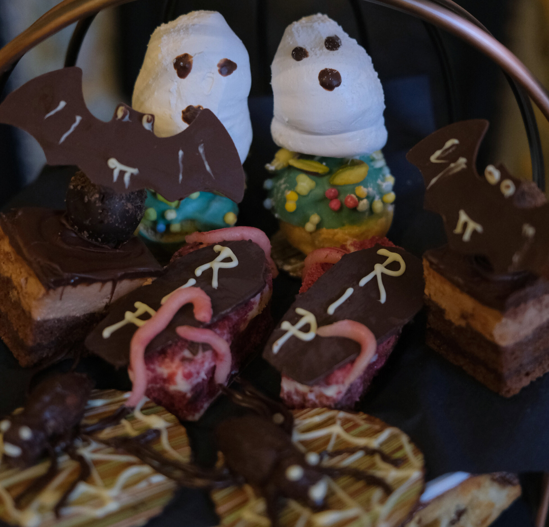 A tiered tray of Halloween-themed desserts including ghost-shaped pastries, chocolate bat decorations, gravestone brownies with 'RIP' written in icing, and cookies with chocolate spiders.