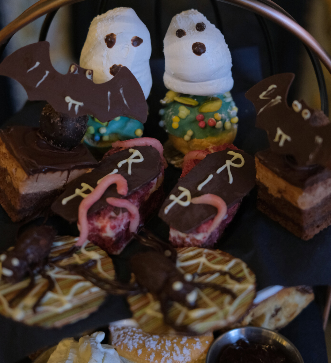 A tiered tray of Halloween-themed desserts including ghost-shaped pastries, chocolate bat decorations, gravestone brownies with 'RIP' written in icing, and cookies with chocolate spiders.