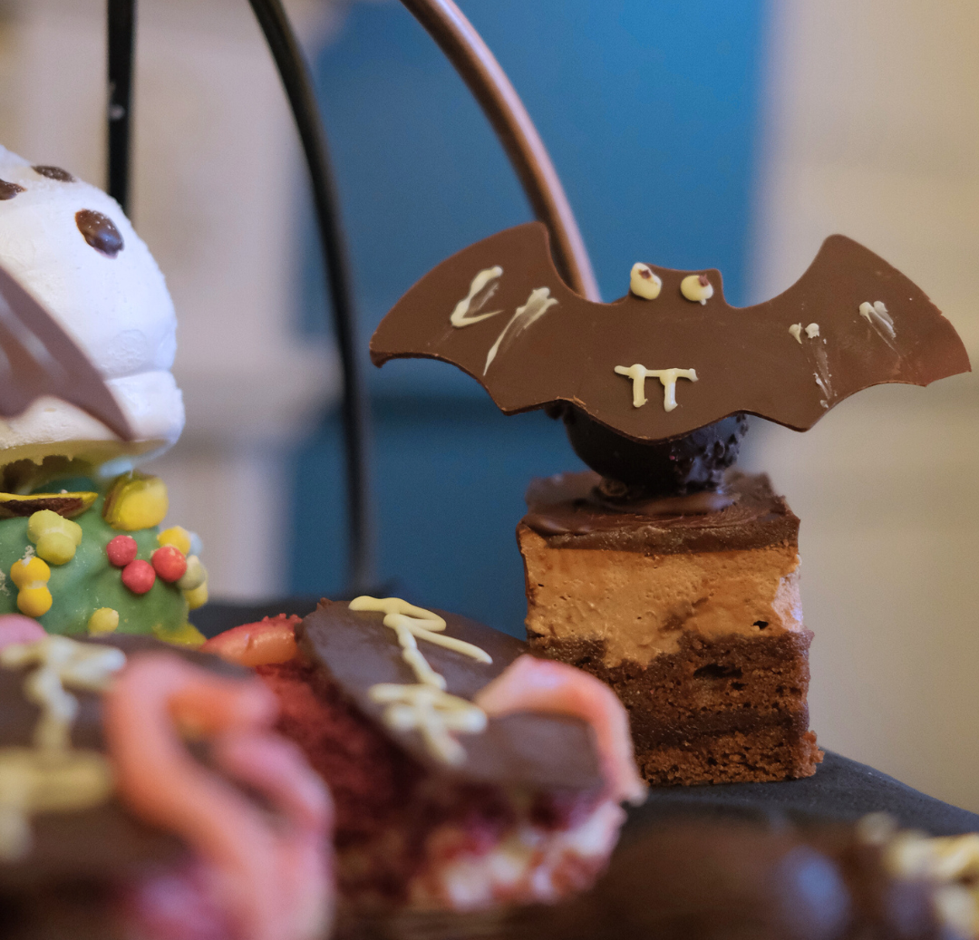 Halloween-themed desserts with a chocolate bat perched on a layered square cake, surrounded by other decorated treats