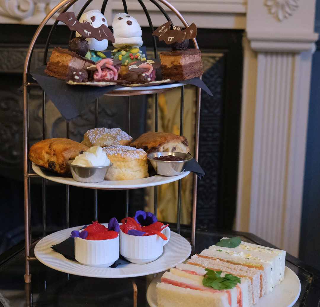A Halloween-themed afternoon tea set featuring sandwiches, scones with cream and jam, and decorated Halloween pastries with ghost and bat designs.