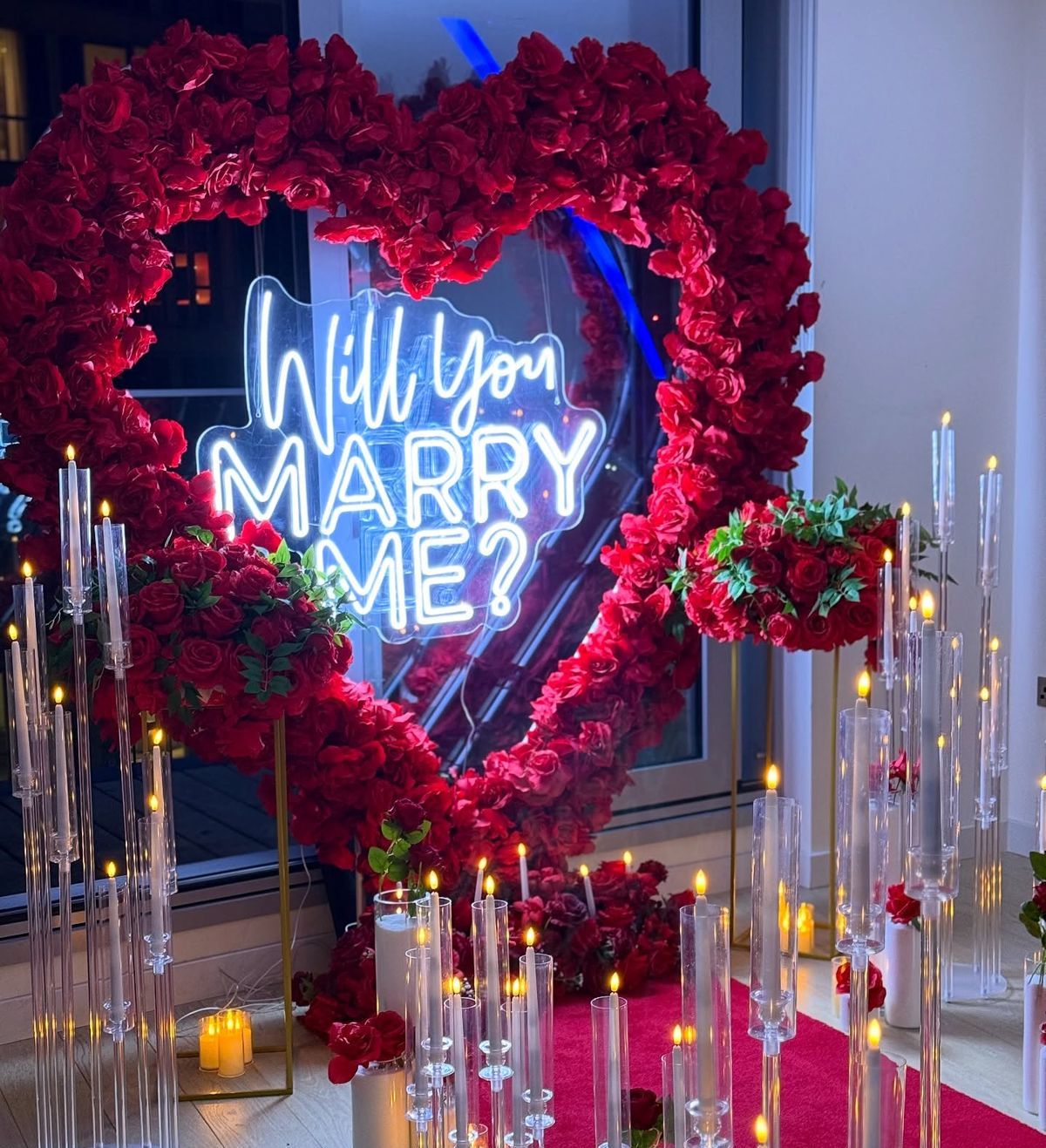 Romantic proposal setup with red roses and a neon sign that reads 'Will You Marry Me?'