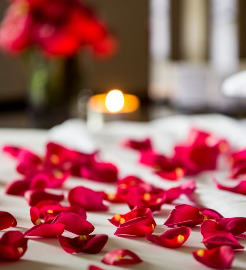 Rose petals scattered on a bed with a candle and flowers in the background