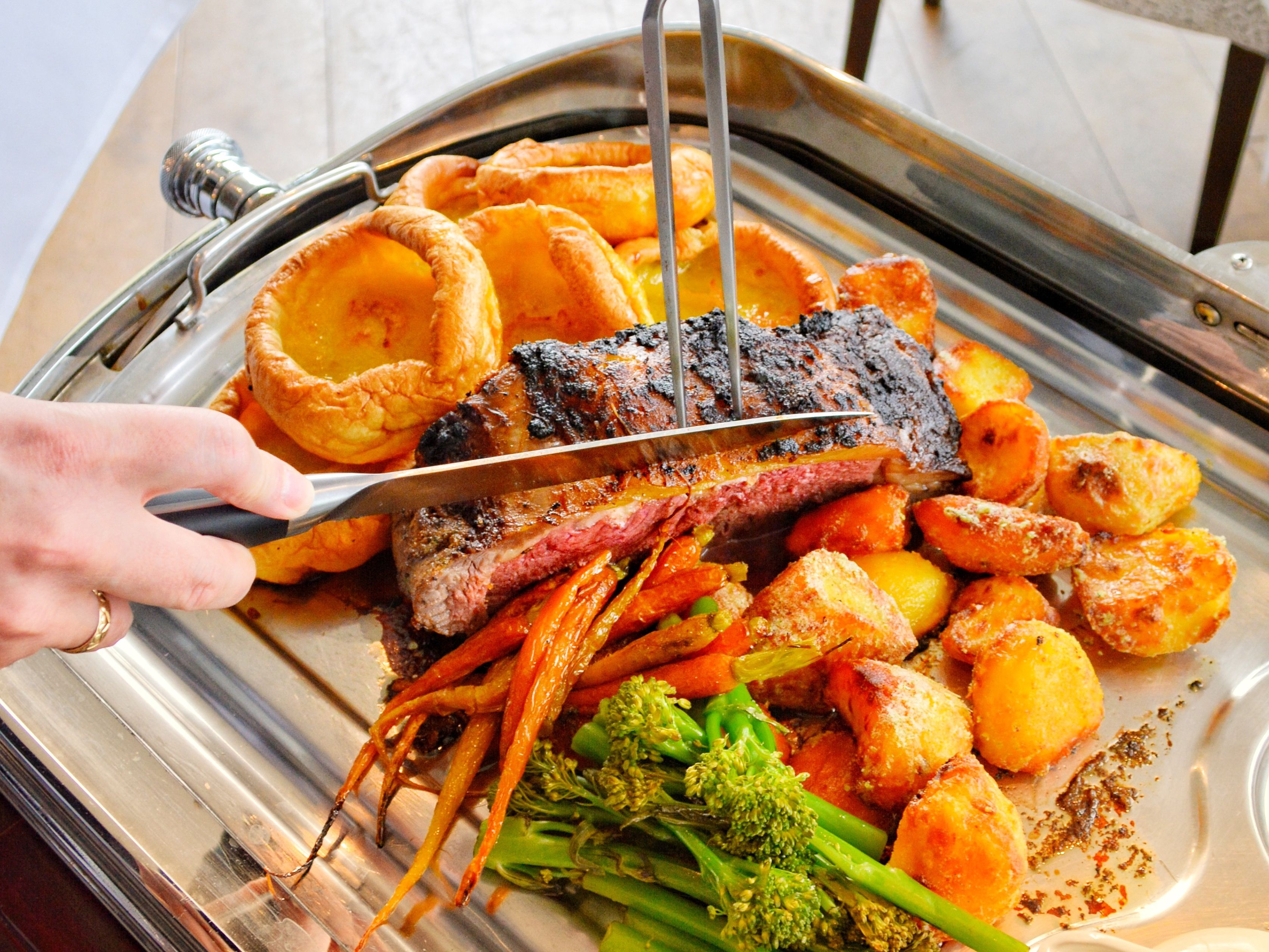 Person carving a roast beef with roast potatoes, Yorkshire puddings, carrots, and broccoli on a tray