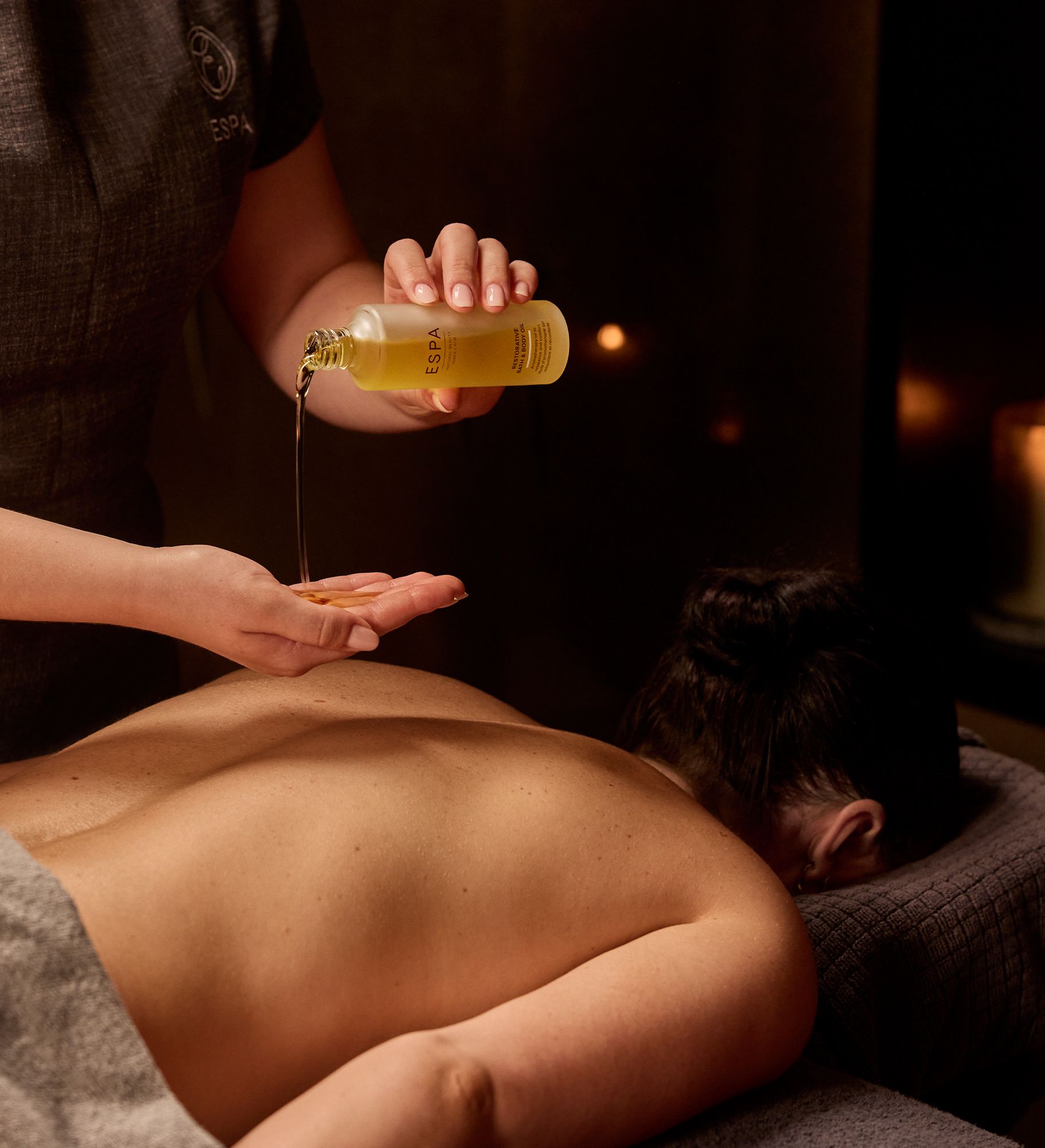 Massage therapist pouring oil onto their hand during a spa treatment with a candle burning in the background.