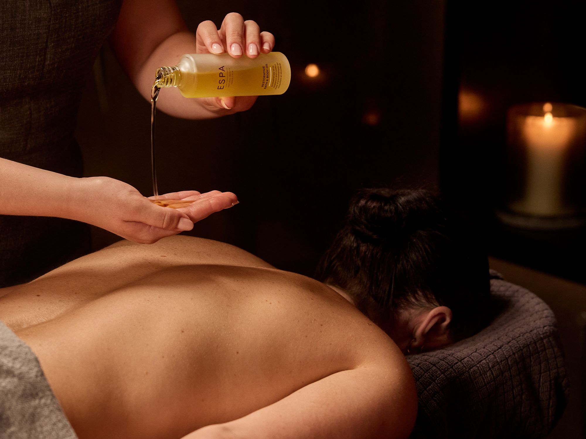 Massage therapist pouring oil onto their hand during a spa treatment with a candle burning in the background.