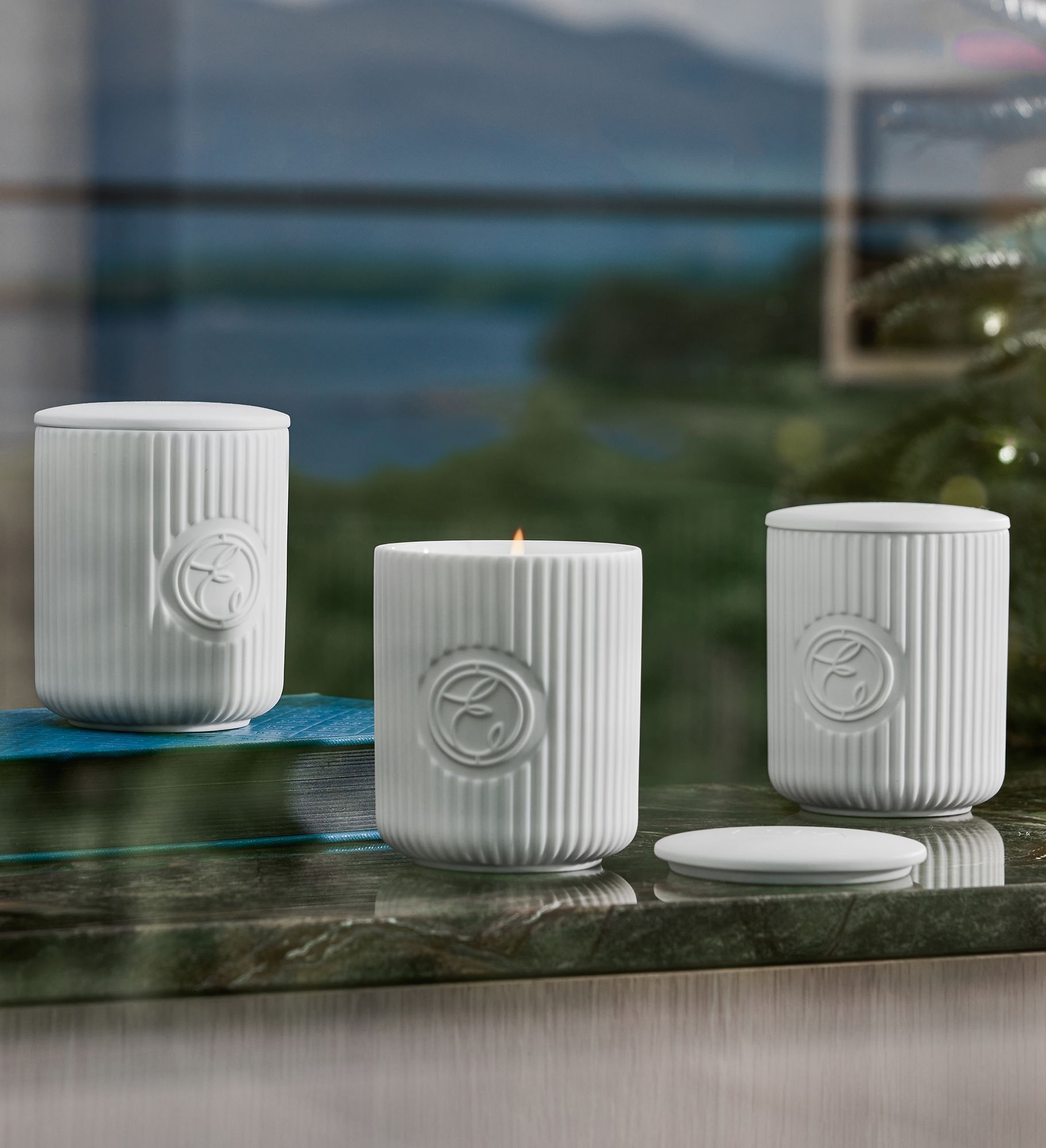 Three white ribbed ceramic candles, one lit, on a marble surface with a scenic background.