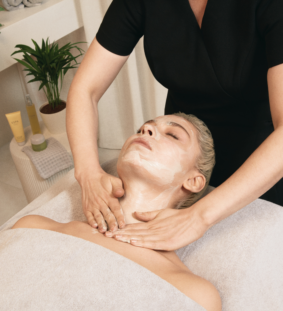 Person receiving a facial treatment at a spa, with a mask applied to the face and a therapist massaging their neck.