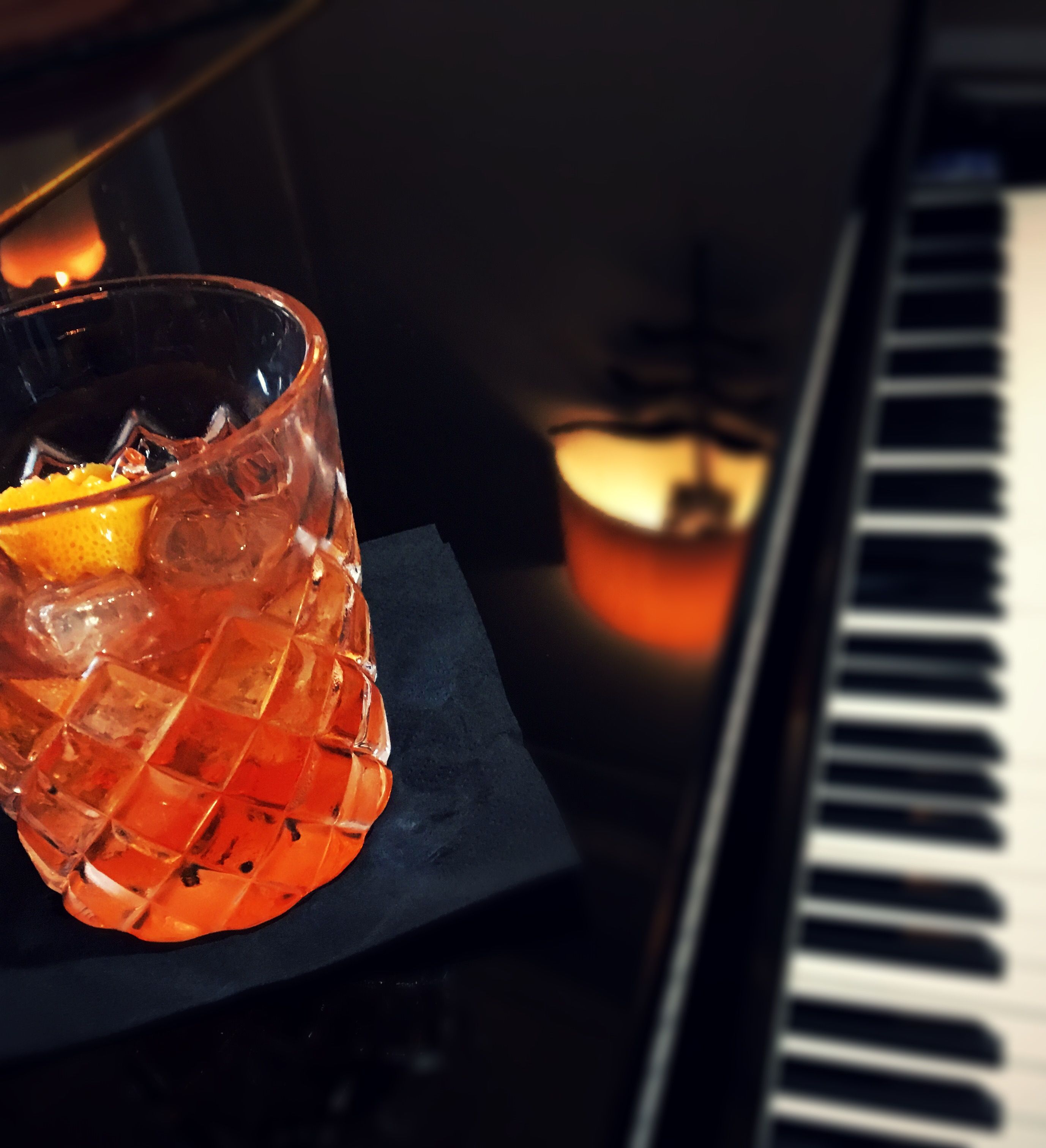 A glass of cocktail with an orange peel garnish on a black napkin, beside a piano keyboard.