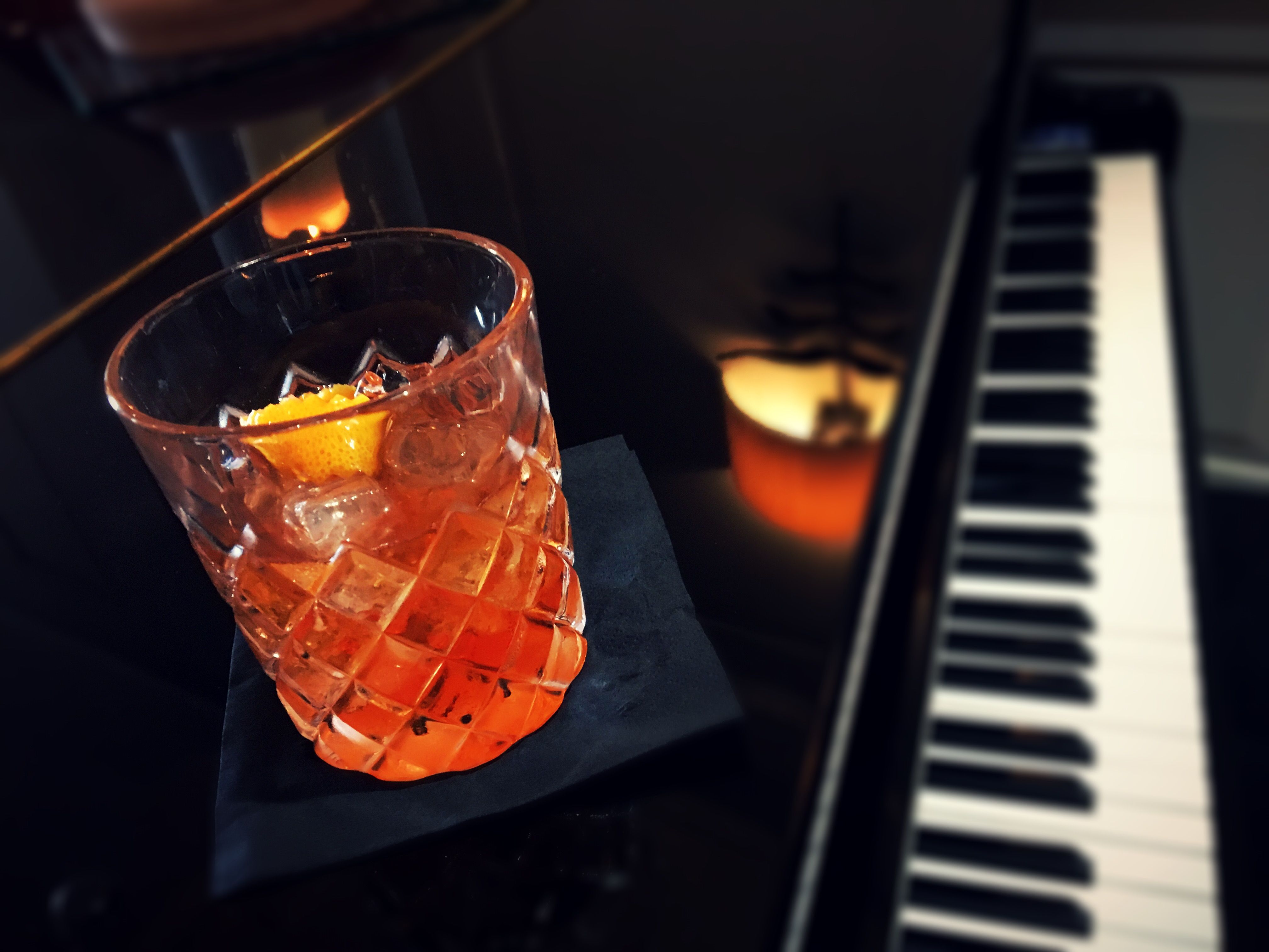 A glass of cocktail with an orange peel garnish on a black napkin, beside a piano keyboard.