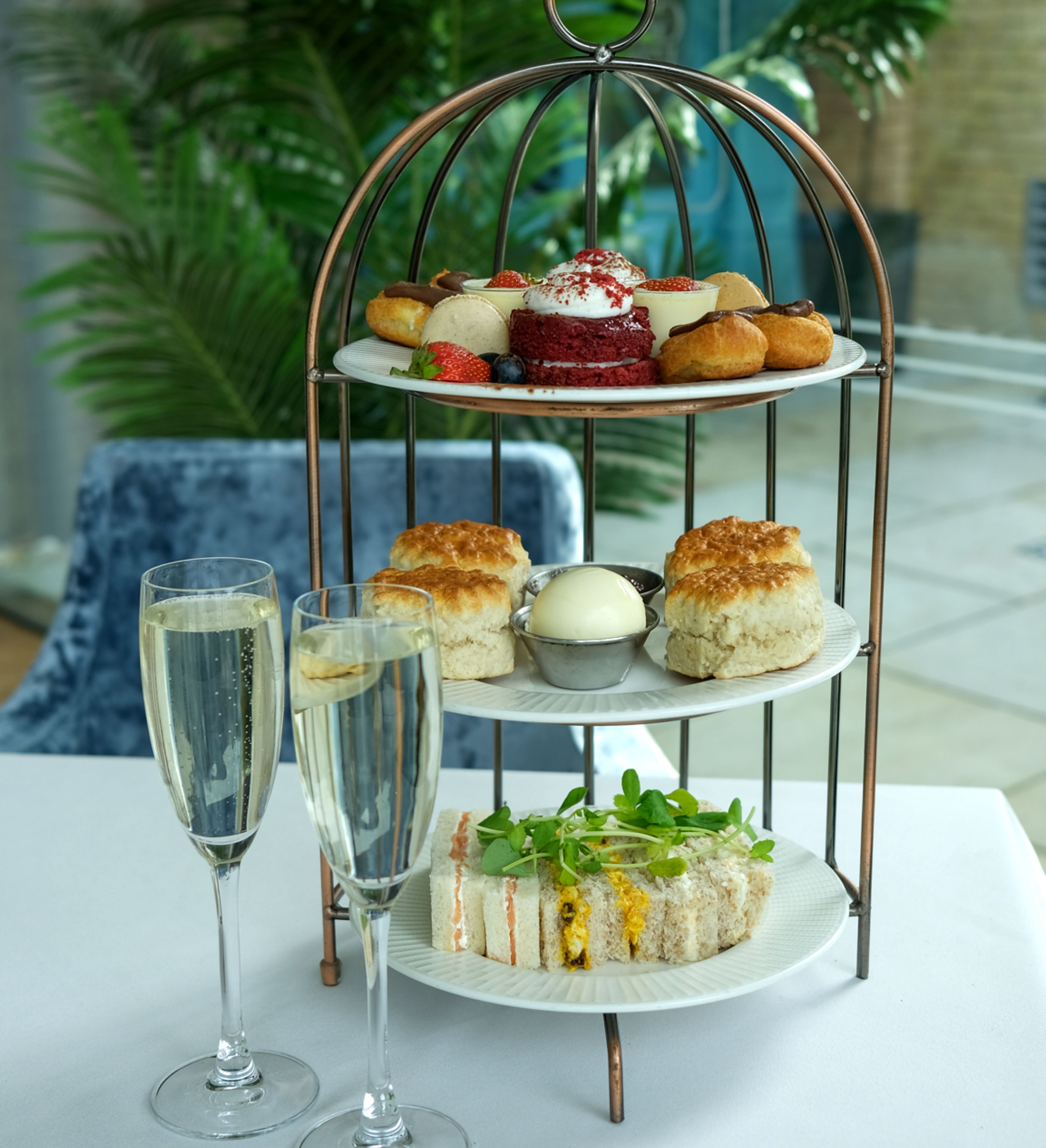 Three-tiered stand with assorted afternoon tea treats and two glasses of champagne on a white tablecloth.