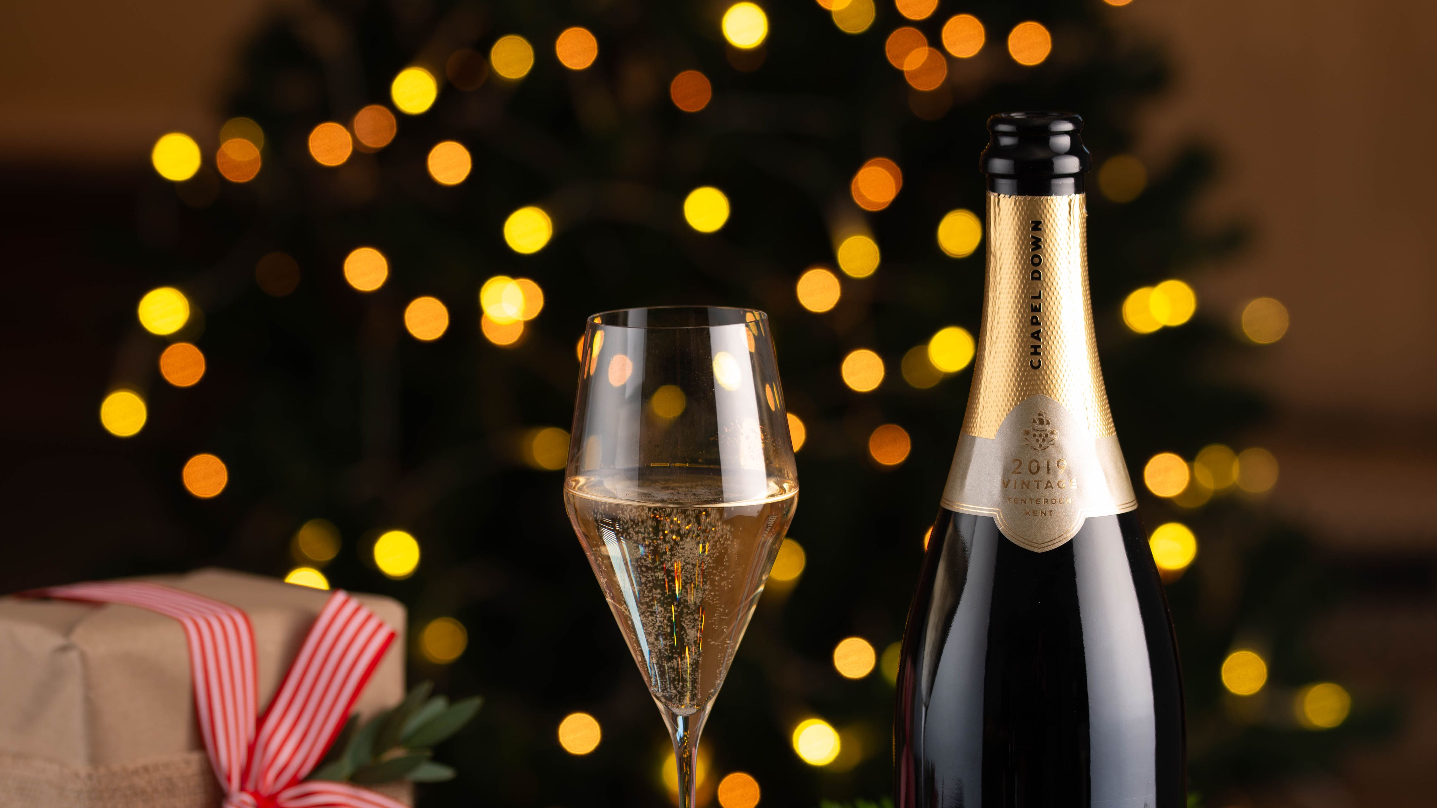 Chapel Down Brut Reserve 2019 full bottle with a filled wine glass, festive cheese platter, and a holiday gift in front of a blurred Christmas tree with lights.