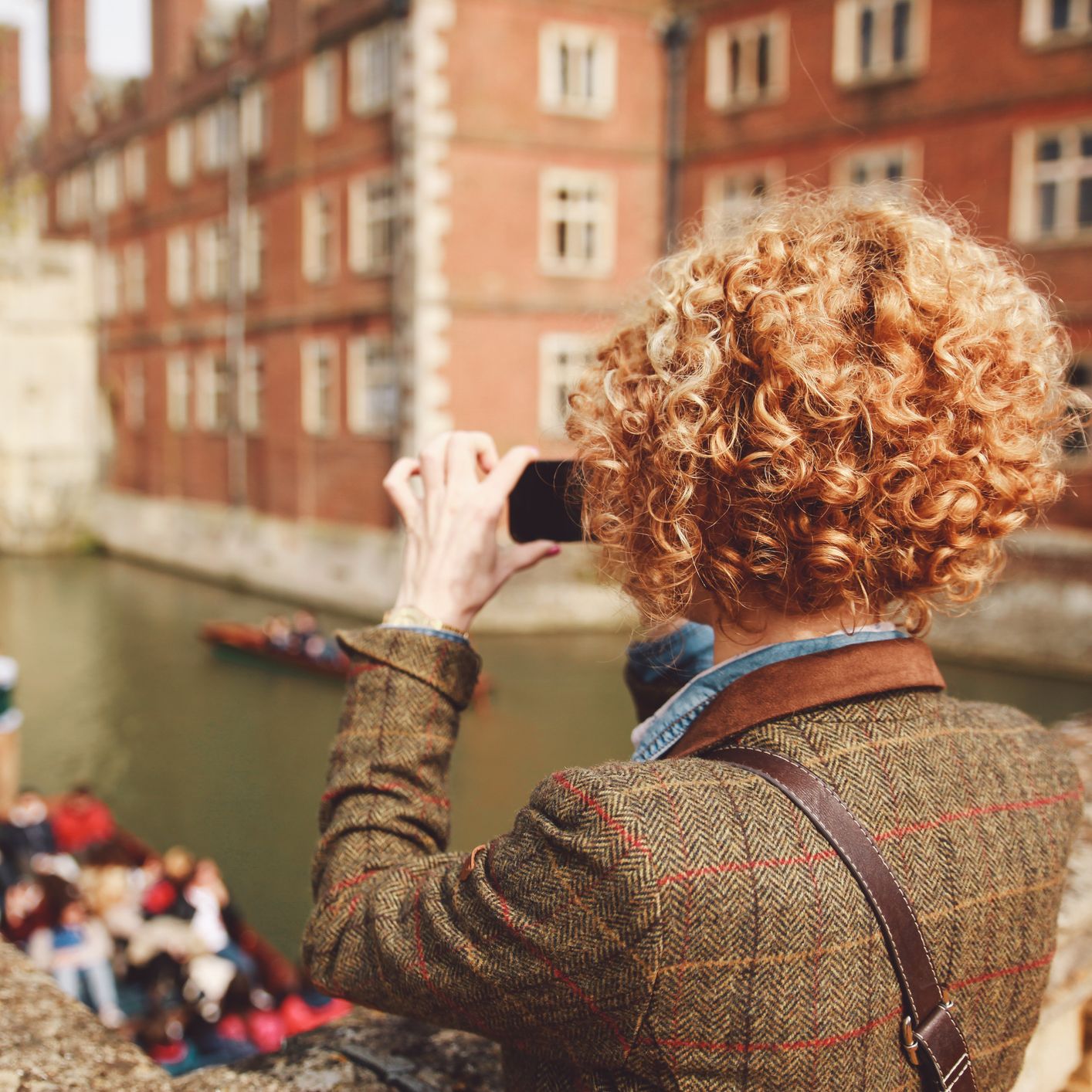 Person with curly hair photographs punts on the River Cam in Cambridge, standing on a stone bridge with historic college buildings in the background.