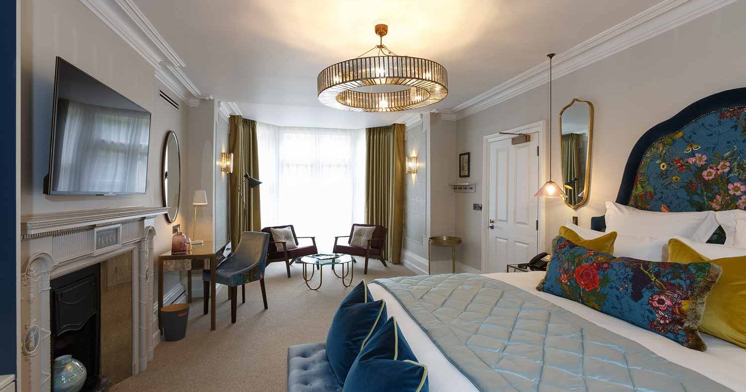 The Delphinium feature room at Gonville Hotel, with a floral headboard, velvet accents, chandelier lighting, seating area, desk, and fireplace.