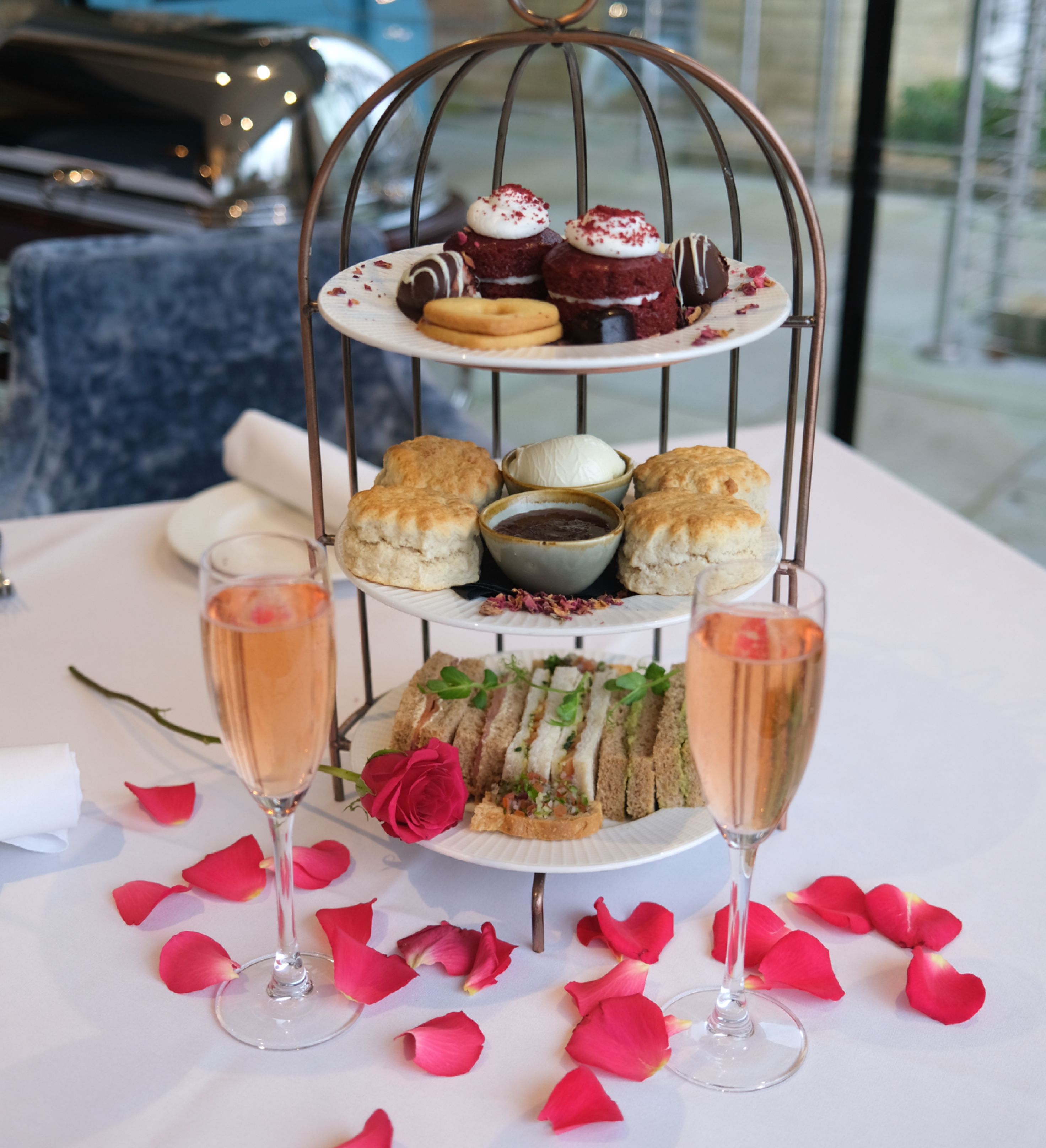 Three-tiered afternoon tea stand with cakes, scones, sandwiches, and two glasses of sparkling rosé on a table decorated with pink rose petals.