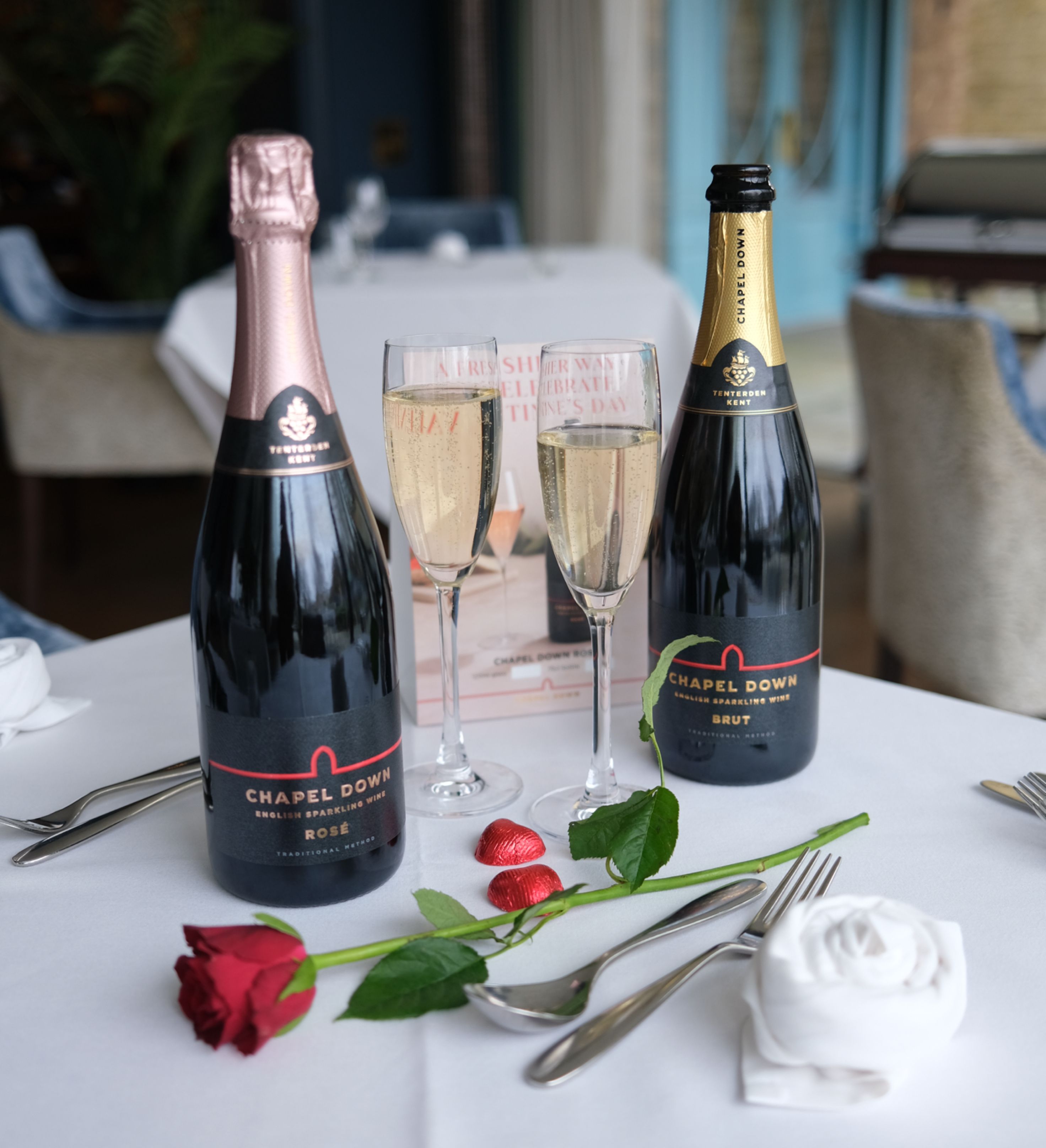 Two bottles of Chapel Down sparkling wine with two filled glasses, a red rose, and heart-shaped chocolates on a table set for a romantic occasion.