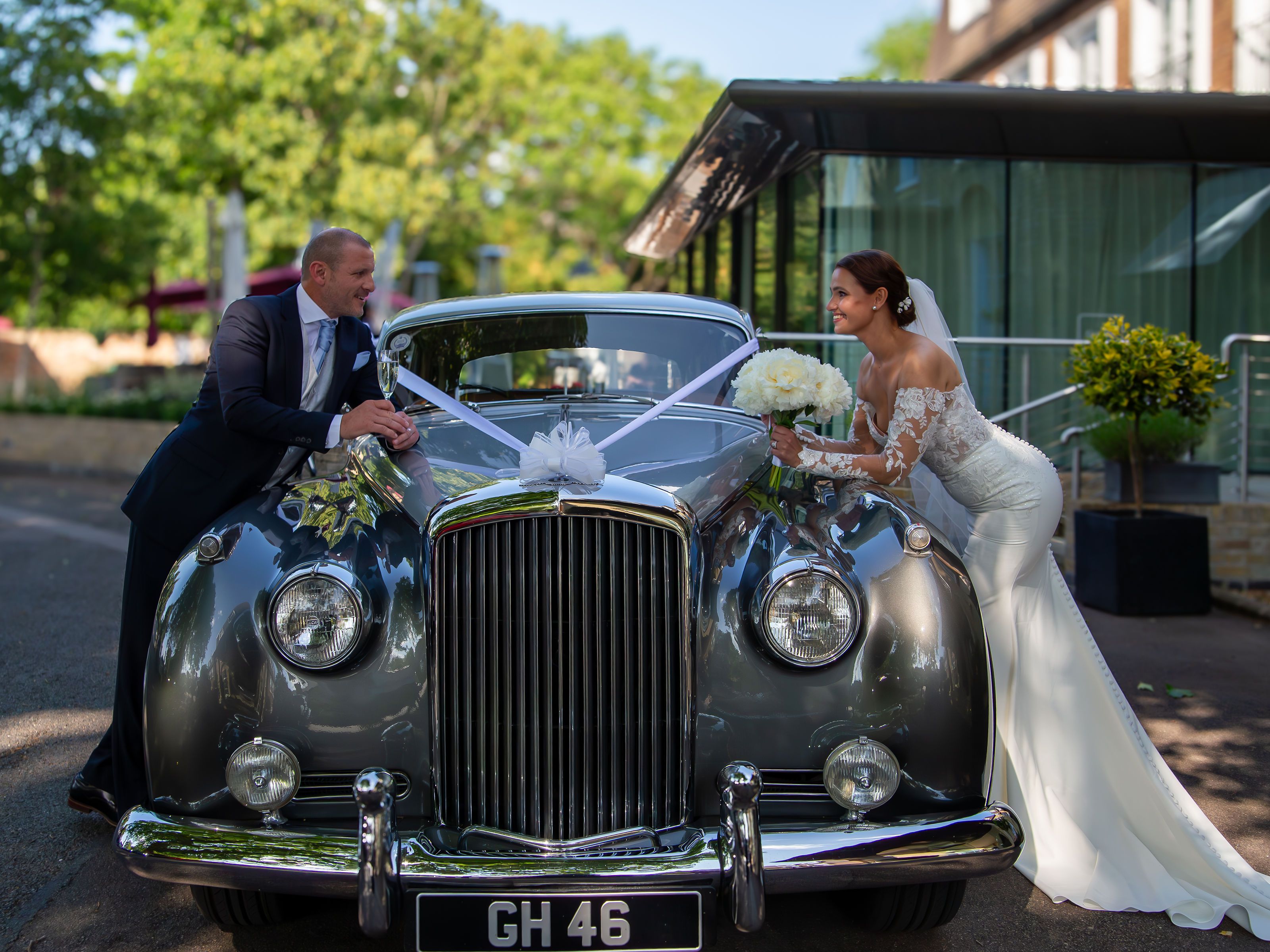 Bride and groom smiling at each other over a classic car decorated with wedding ribbons and flowers