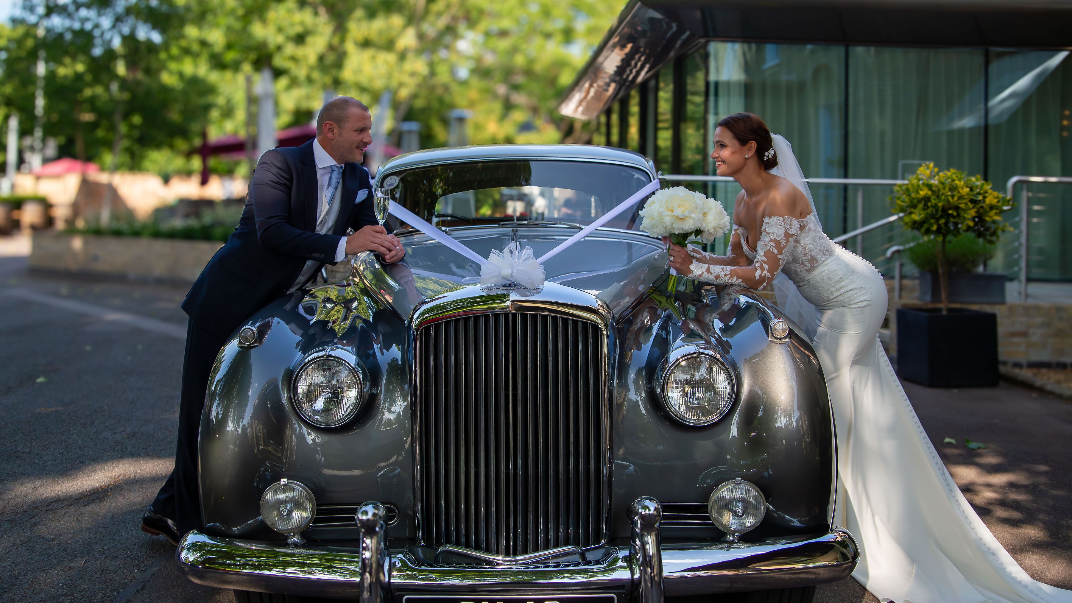 Bride and groom smiling at each other over a classic car decorated with wedding ribbons and flowers