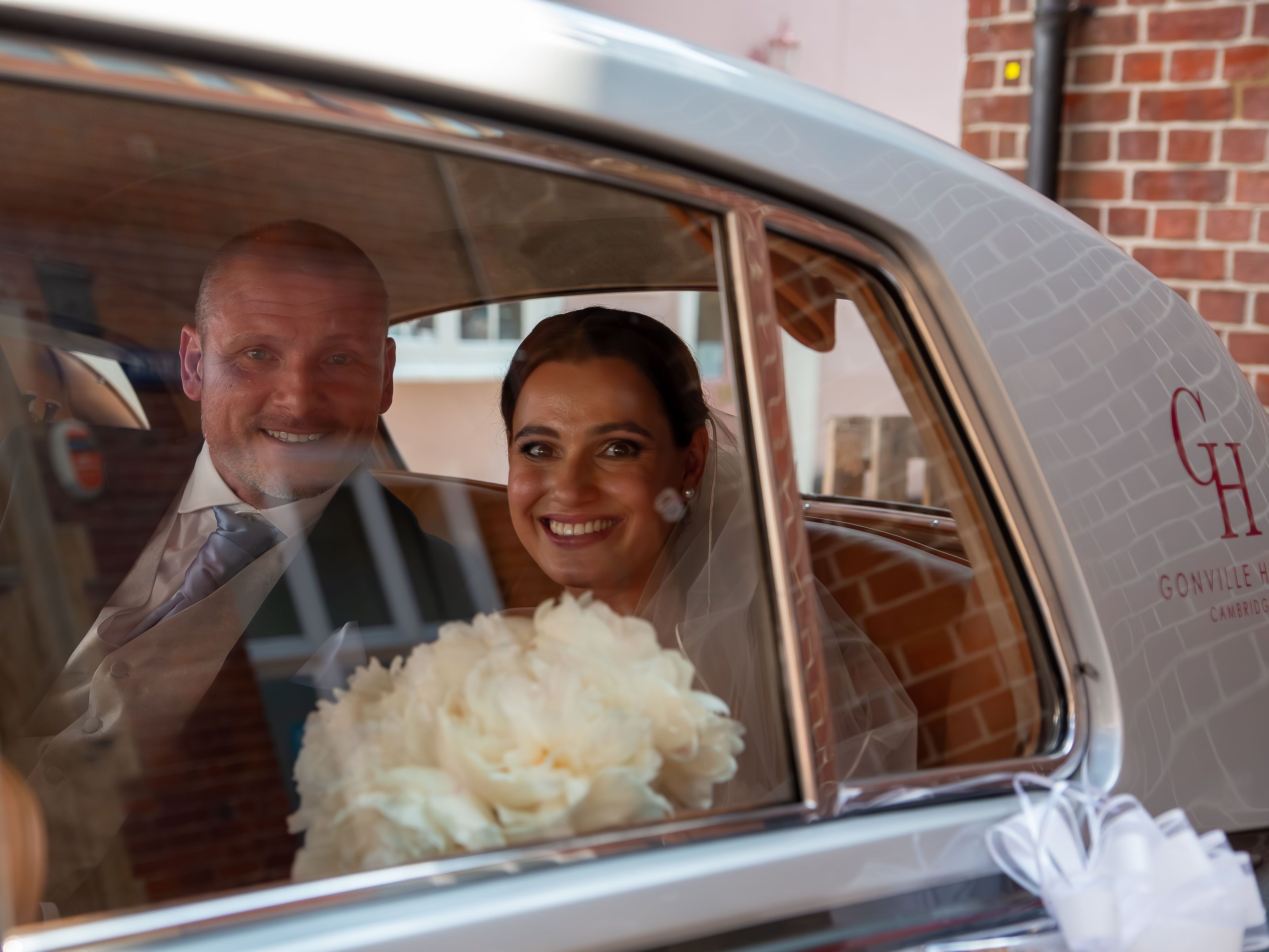 Bride and groom smiling inside a classic car, holding a bouquet of flowers.