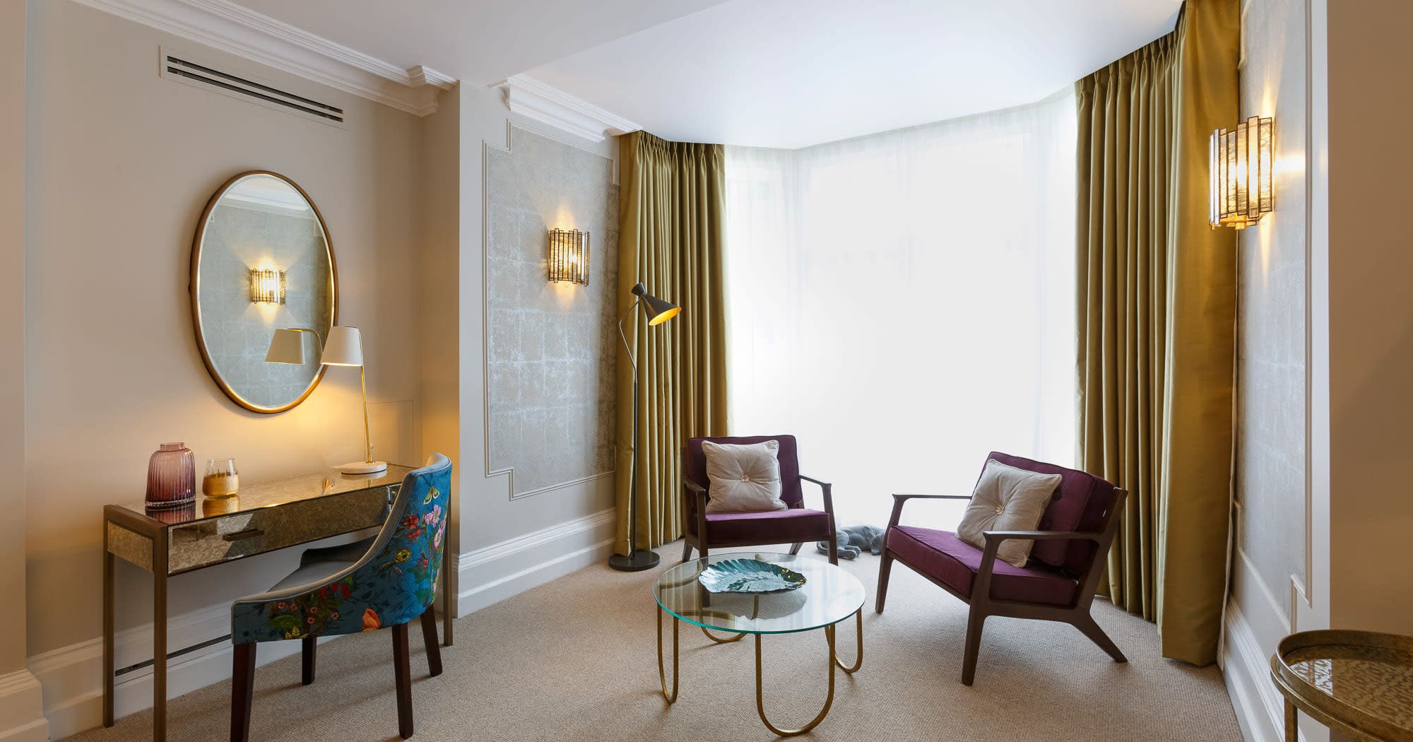 Seating area in the Delphinium feature room at Gonville Hotel, with velvet chairs, a glass coffee table, and a desk with floral-upholstered chair.