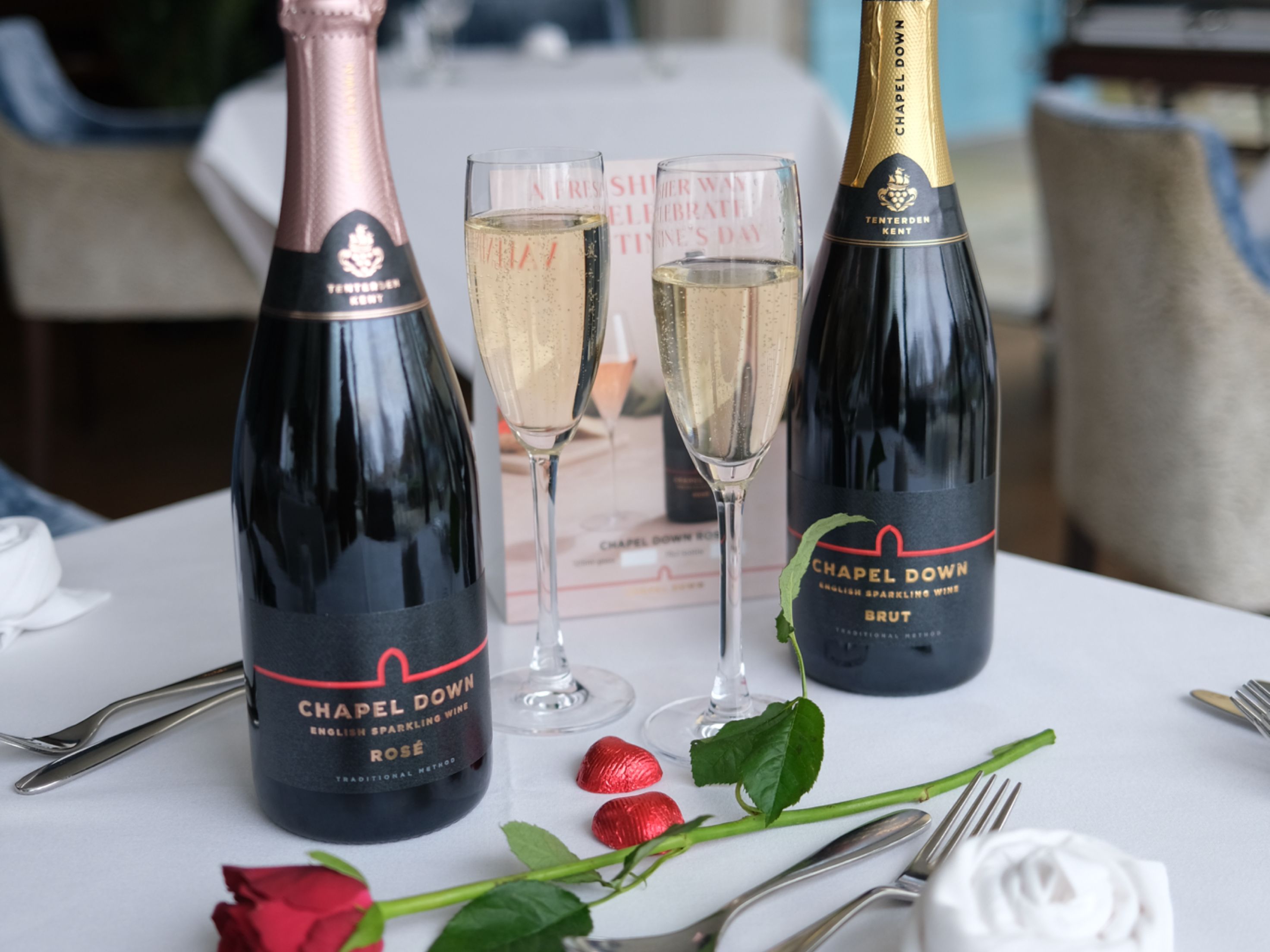 Chapel Down sparkling wine bottles with two filled glasses, a red rose, and heart-shaped chocolates on a restaurant table