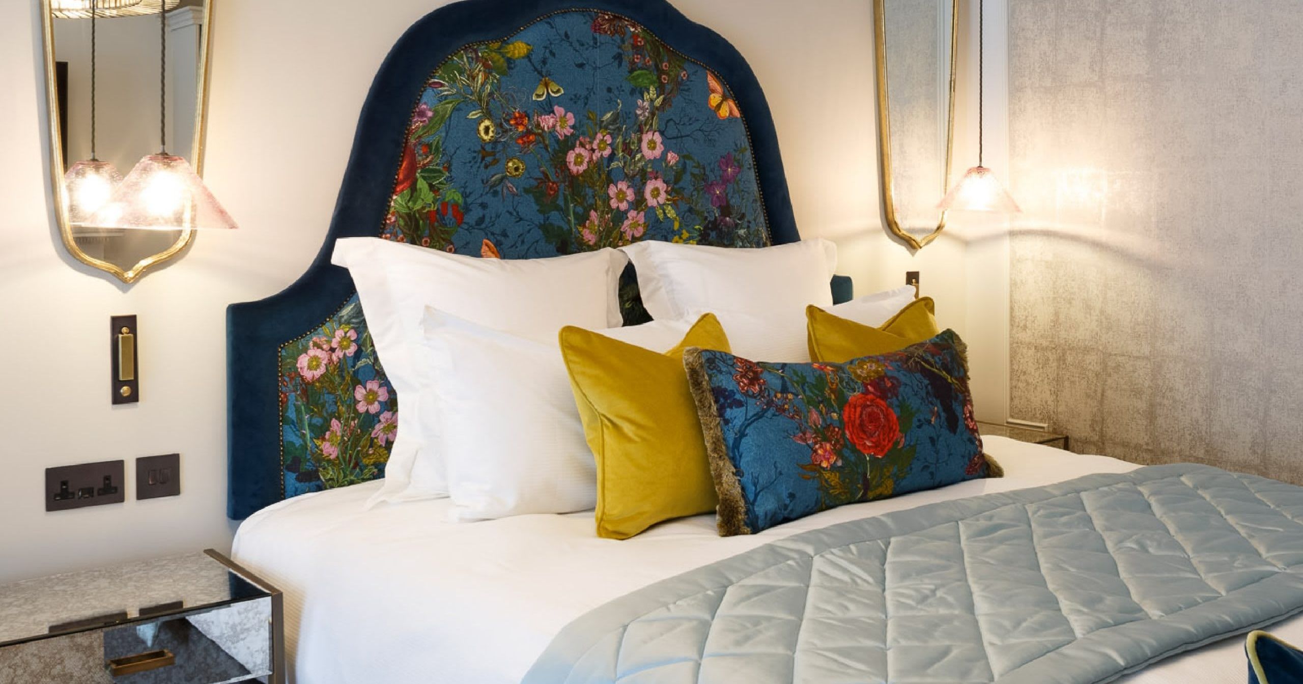 The Delphinium feature room at Gonville Hotel, showcasing a floral blue headboard, vibrant cushions, mirrored bedside tables, and elegant lighting.