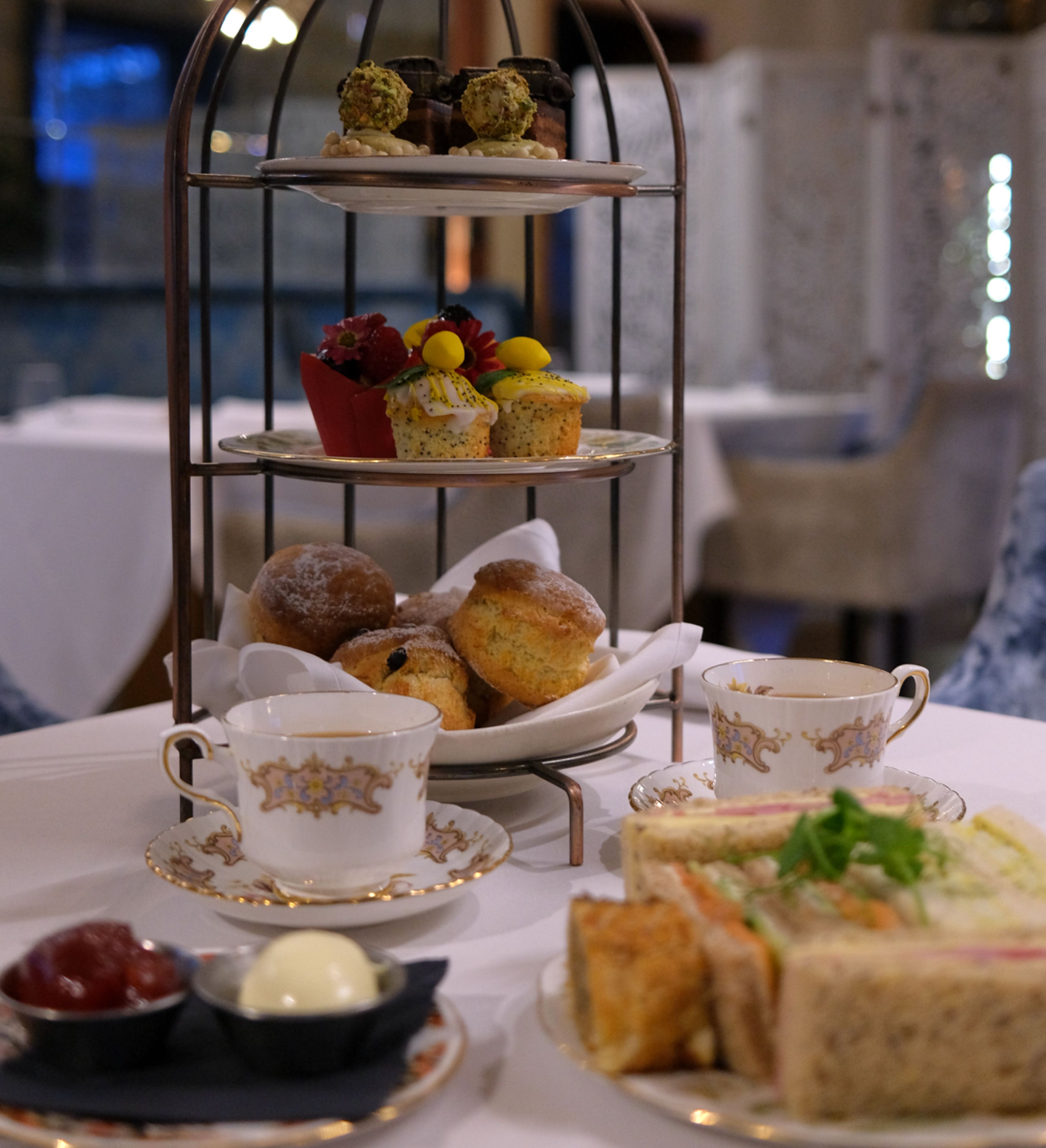 Afternoon tea set with a tiered tray of pastries, scones, and sweets, accompanied by cups of tea and plates of sandwiches.