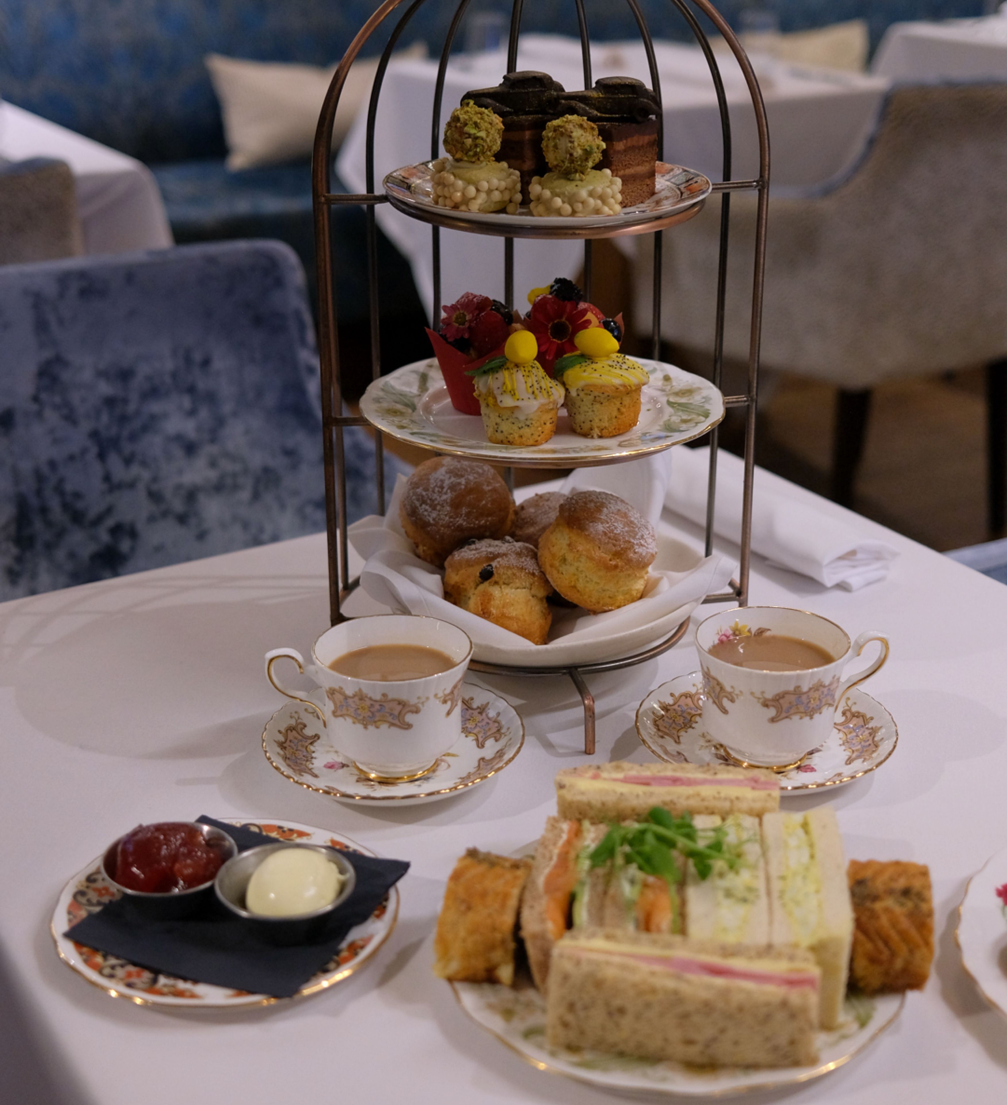 A traditional afternoon tea setup with a three-tiered stand holding pastries, scones, and desserts, surrounded by cups of tea and a plate of assorted finger sandwiches.