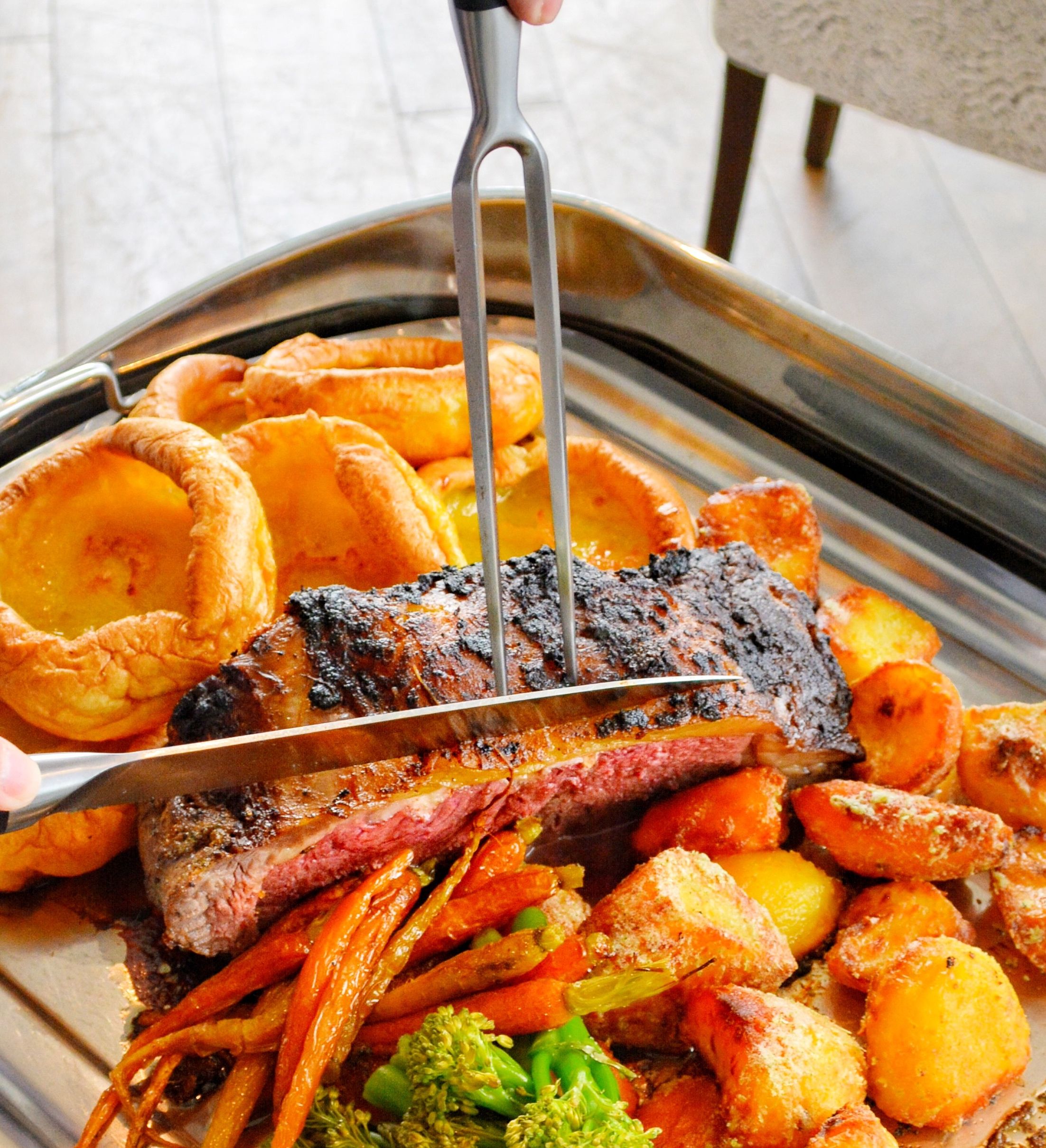 Carving roast beef on a serving tray with Yorkshire puddings, roast potatoes and vegetables