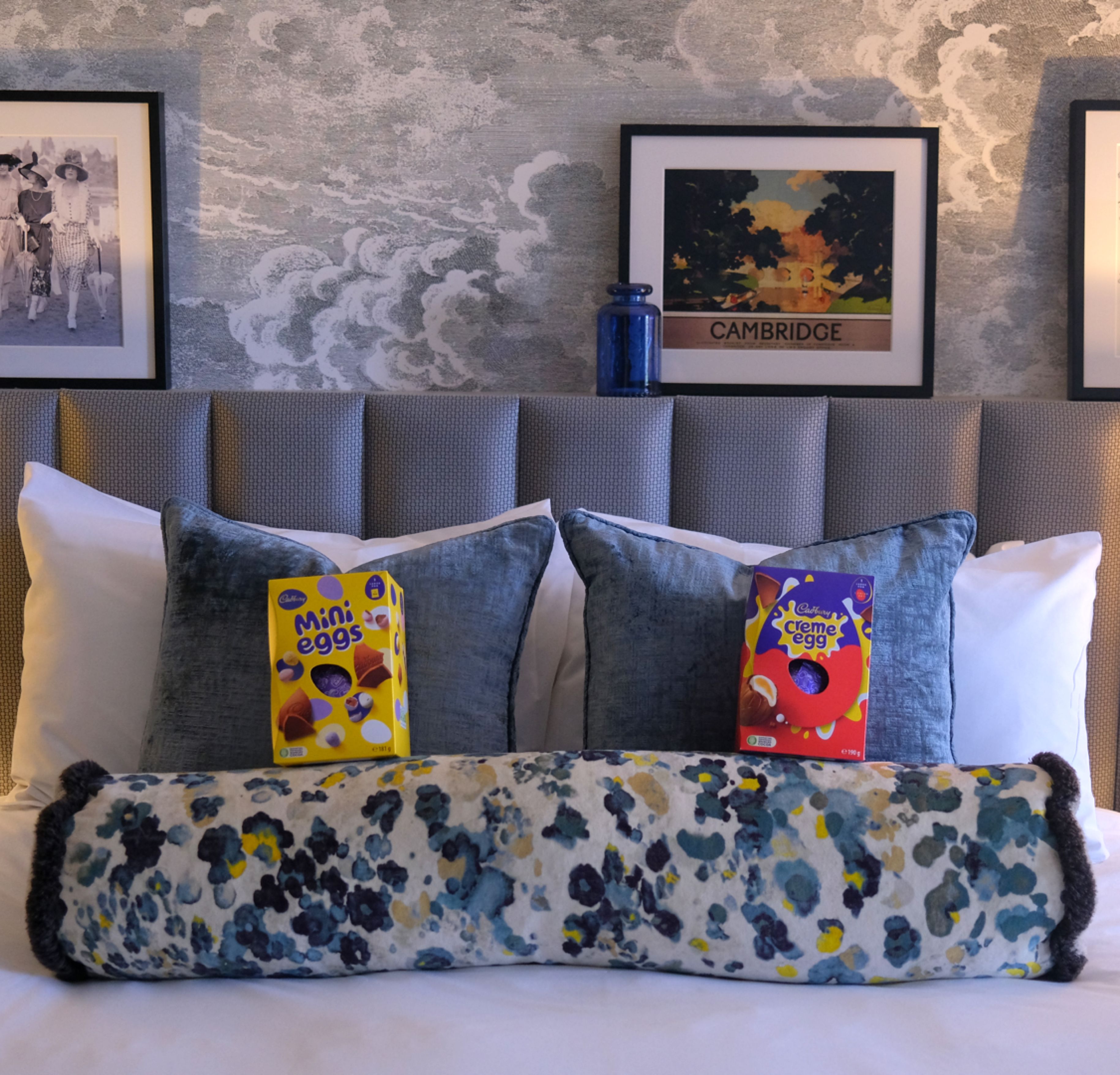 Hotel bed with blue cushions, bedside lamps and framed Cambridge artwork, with Easter chocolate eggs on the pillows