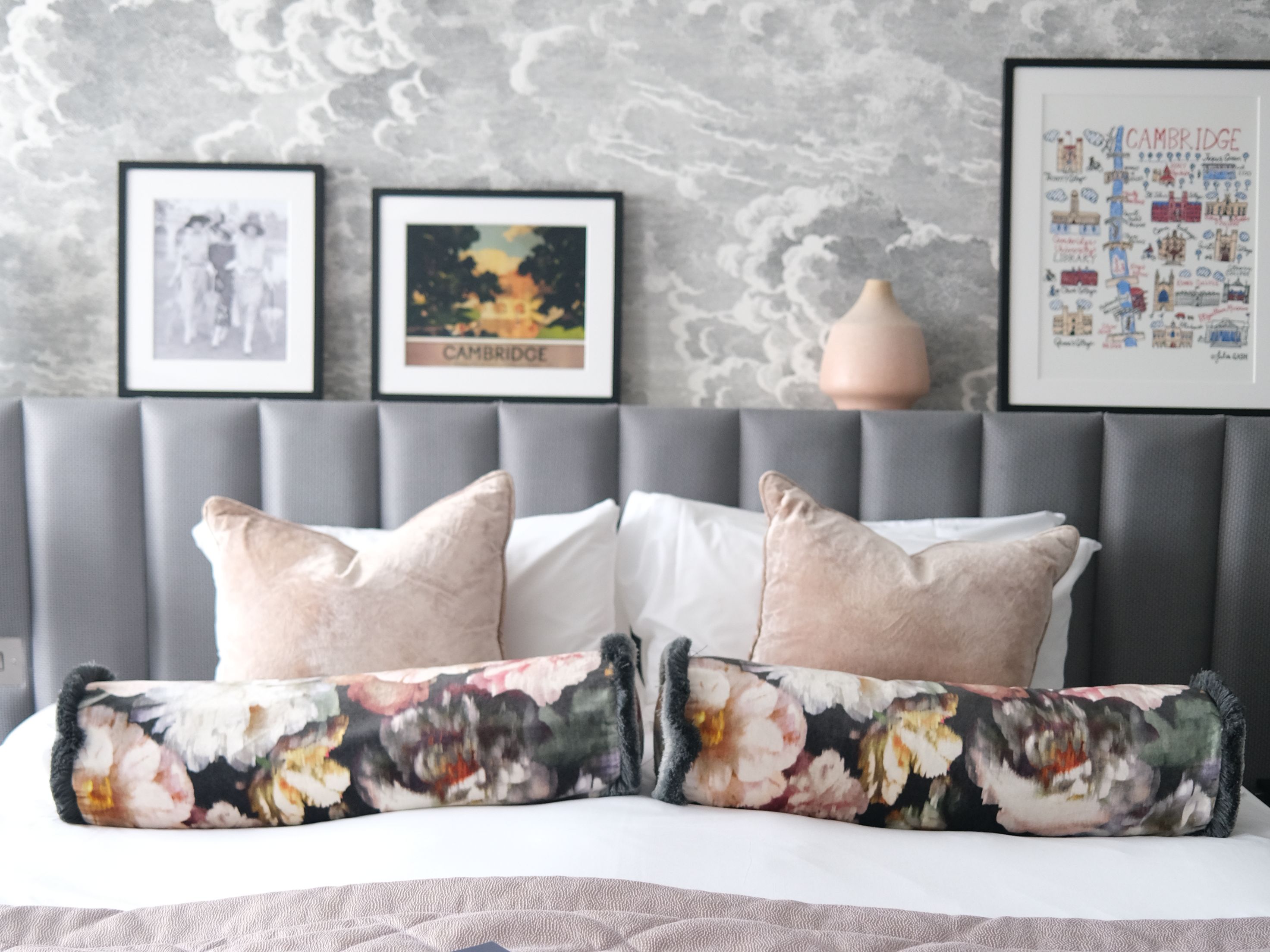 Stylish bedroom with grey upholstered headboard, cloud-pattern wallpaper and framed Cambridge prints above a neatly made bed