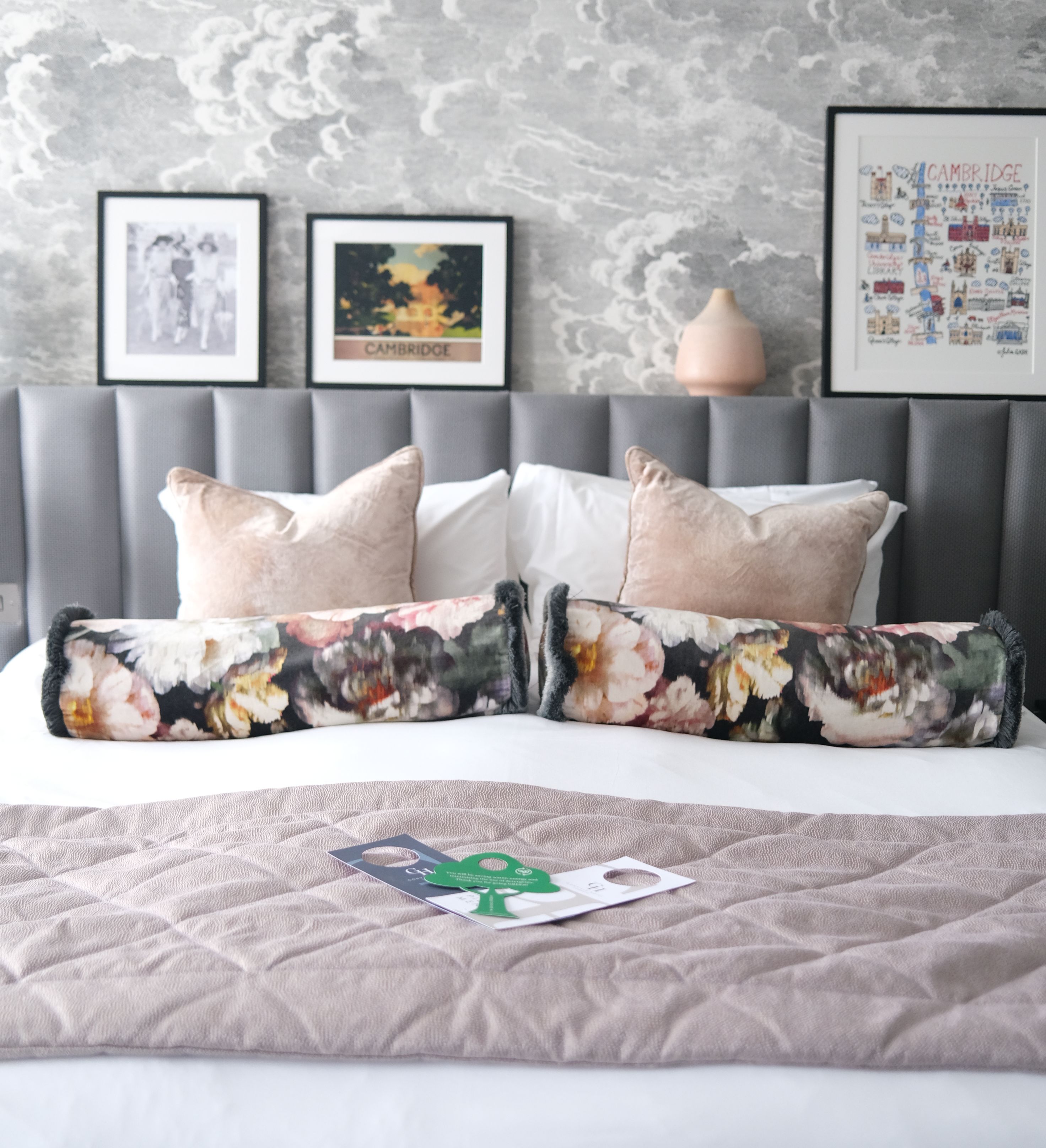 Stylish bedroom with grey upholstered headboard, cloud-pattern wallpaper and framed Cambridge prints above a neatly made bed