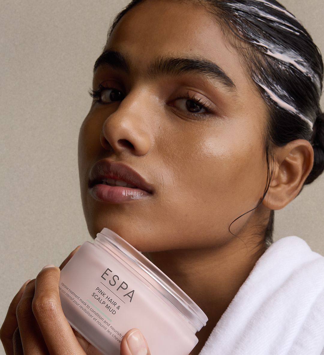 Close-up of a woman in a white robe holding an open ESPA Pink Hair & Scalp Mud jar, with product visible in her hair.