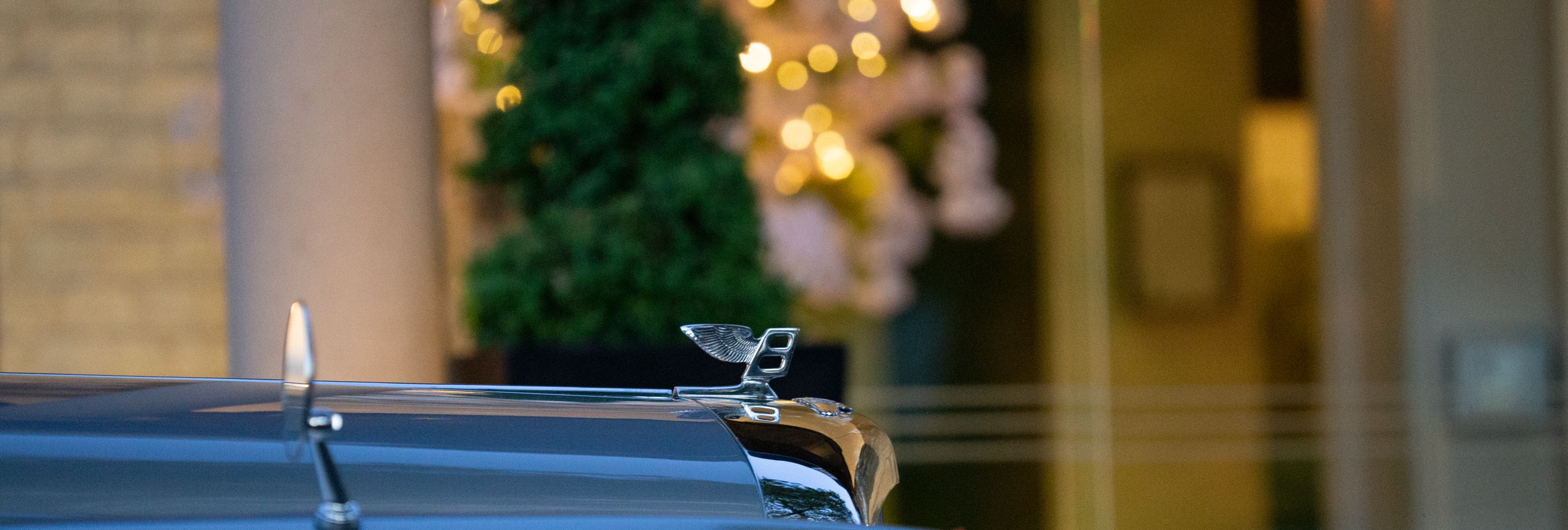 Close-up of a Bentley bonnet mascot on a grey car with warm festive lights blurred in the background