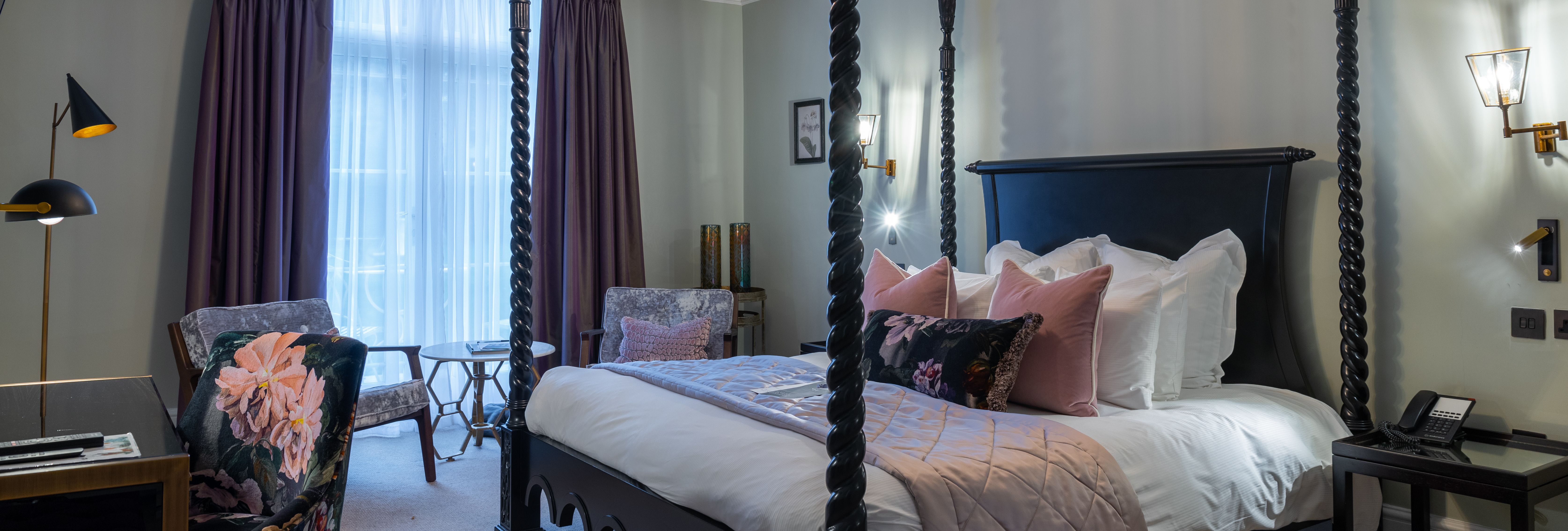 Elegant four-poster bed in a hotel-style bedroom with chandelier, dark curtains and seating area